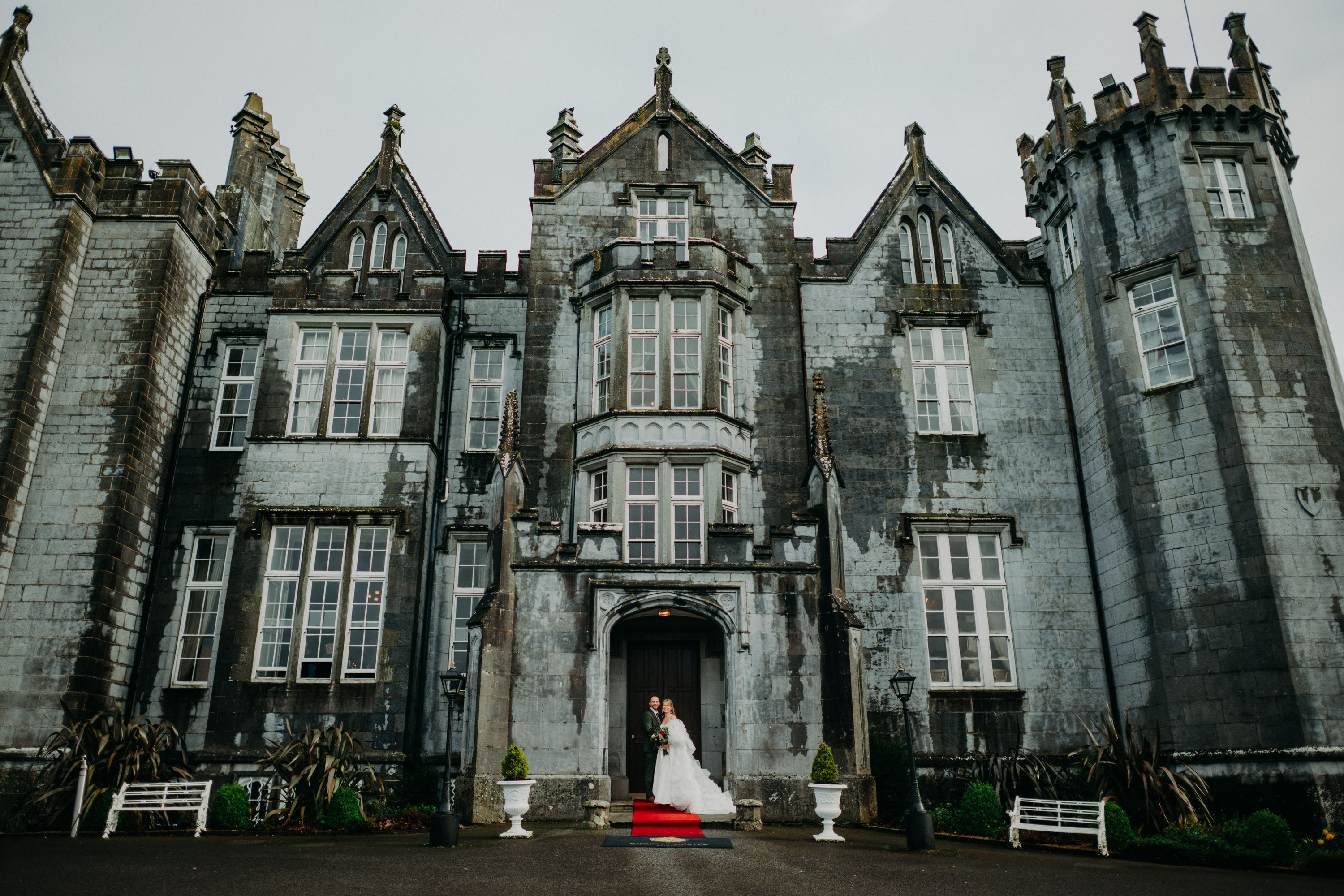 Newlyweds stand on a red carpet before Kinnitty Castle's turreted grey stone facade and Gothic gables in Ireland.