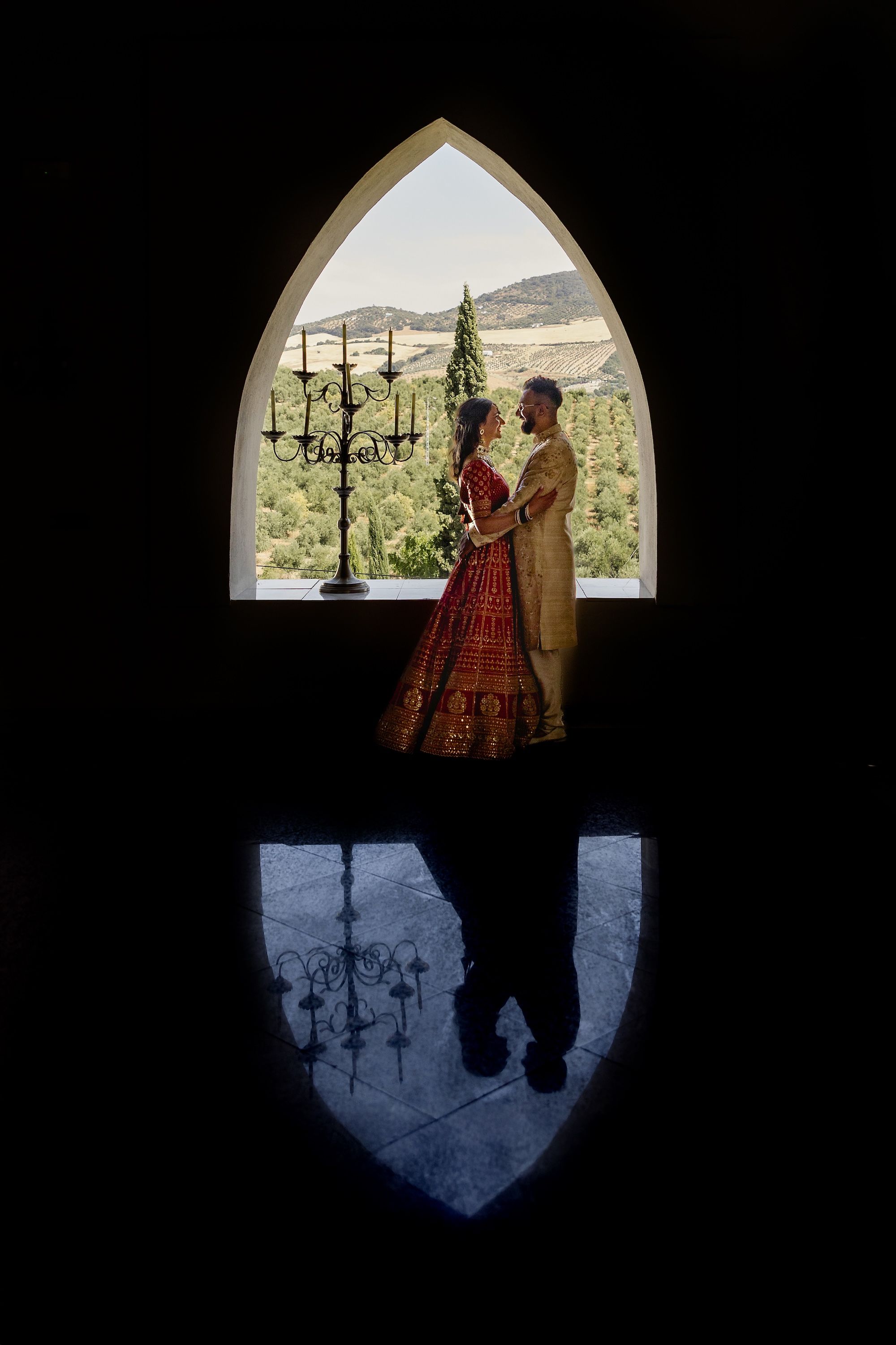 Newlyweds in Hindu wedding attire framed by an open window overlooking vineyards in Cadiz, Spain