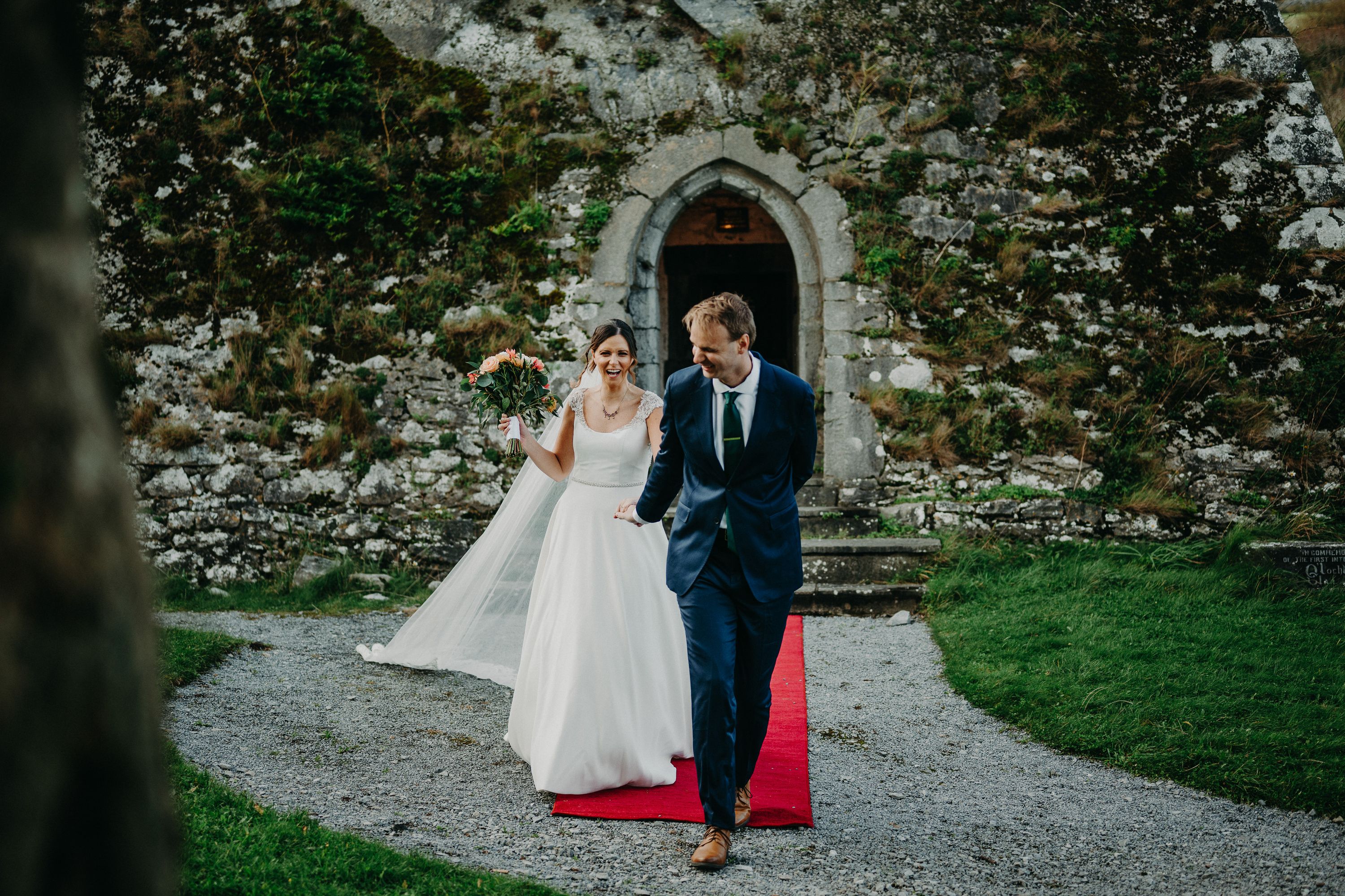 Newlyweds walk hand in hand along a red carpet after their ceremony at a small wedding in Ireland in a medieval castle.