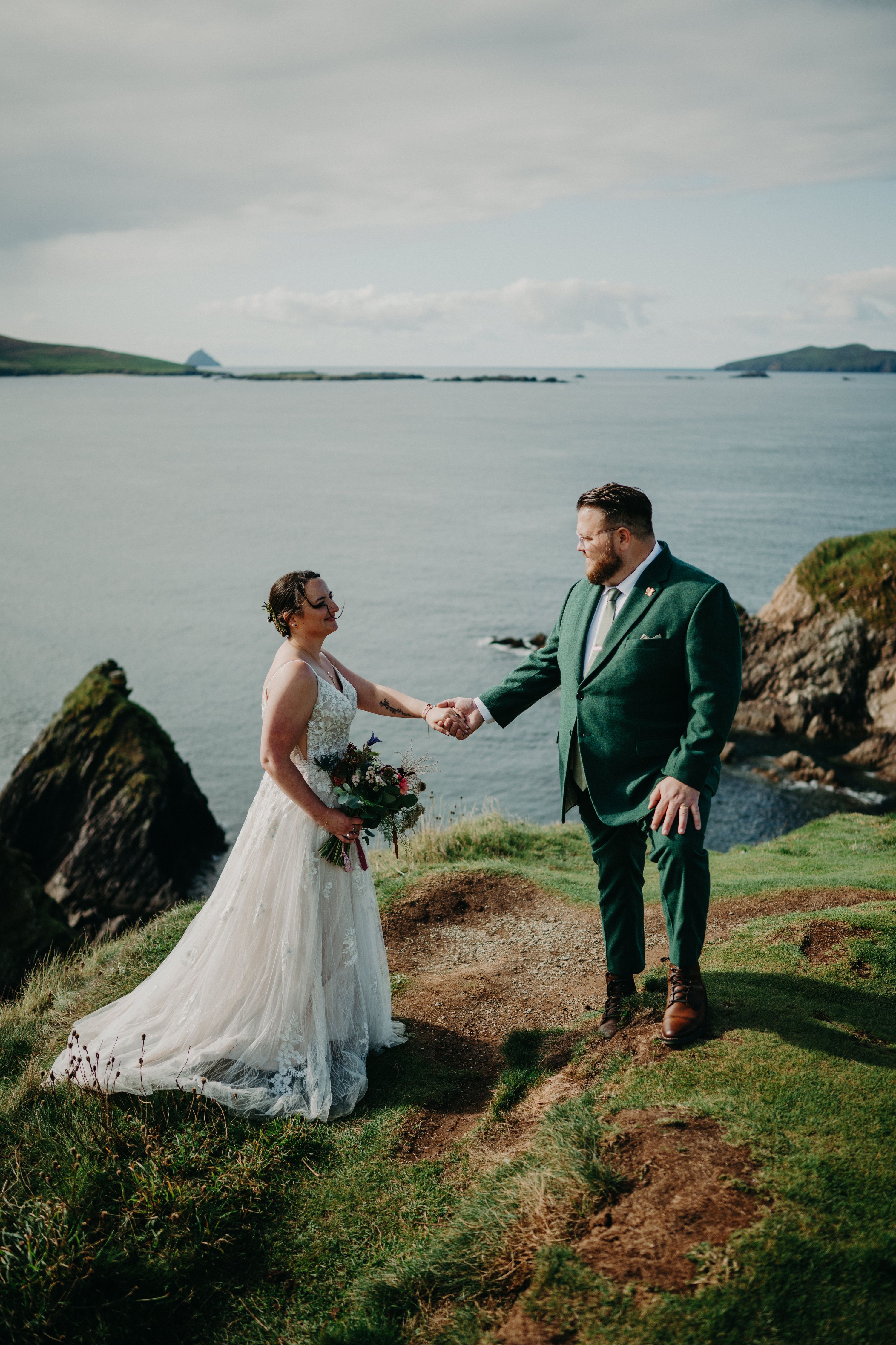 Bride and groom on top of a cliff holding hands with the Dunquin Pier and the Atlantic Ocean in the background