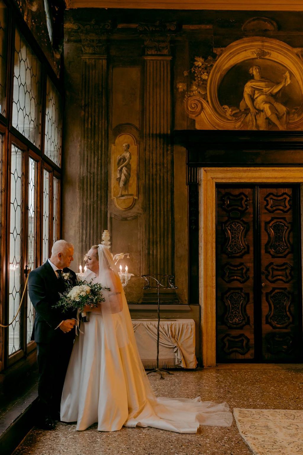 Newlyweds look at each other while standing beside a large palace window during the photoshoot of their vow renewal in Italy