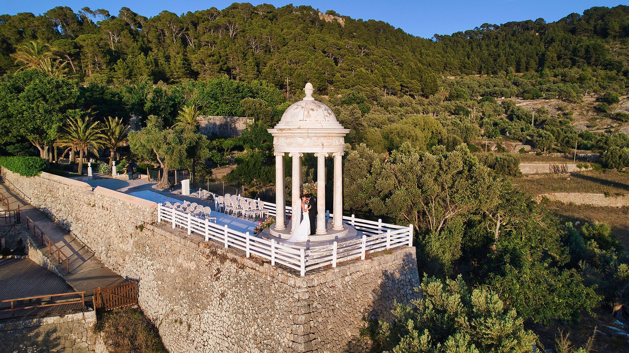 Wide shot photo of a stone gazebo and a ceremony spot perched on a clifftop surrounded by greenery in Mallorca