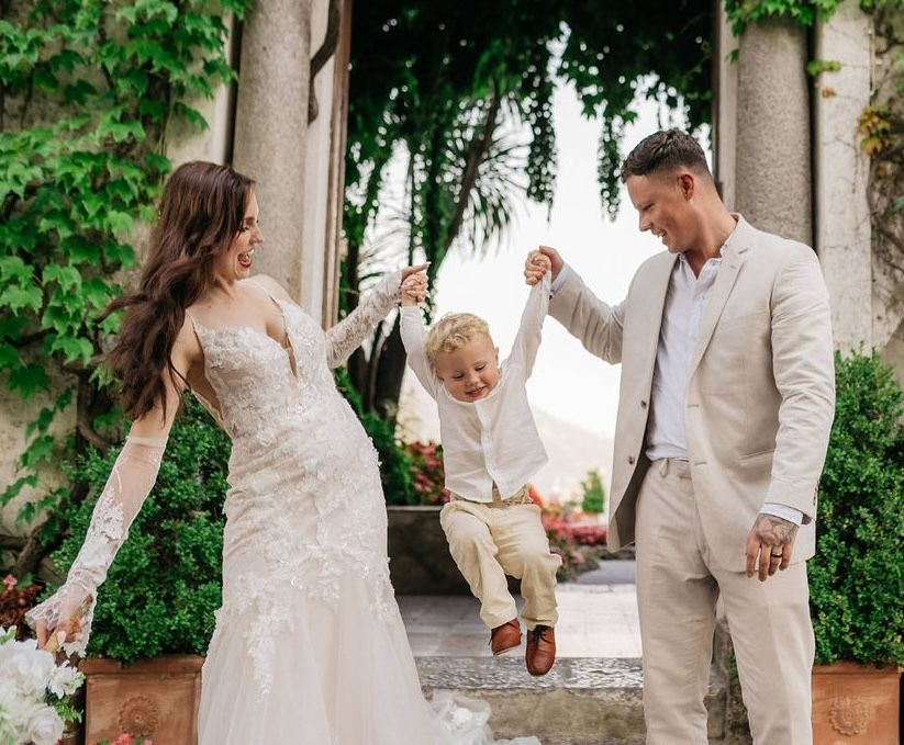 Bride and groom side by side helping each other lift their son who's in the middle, enjoying the activity