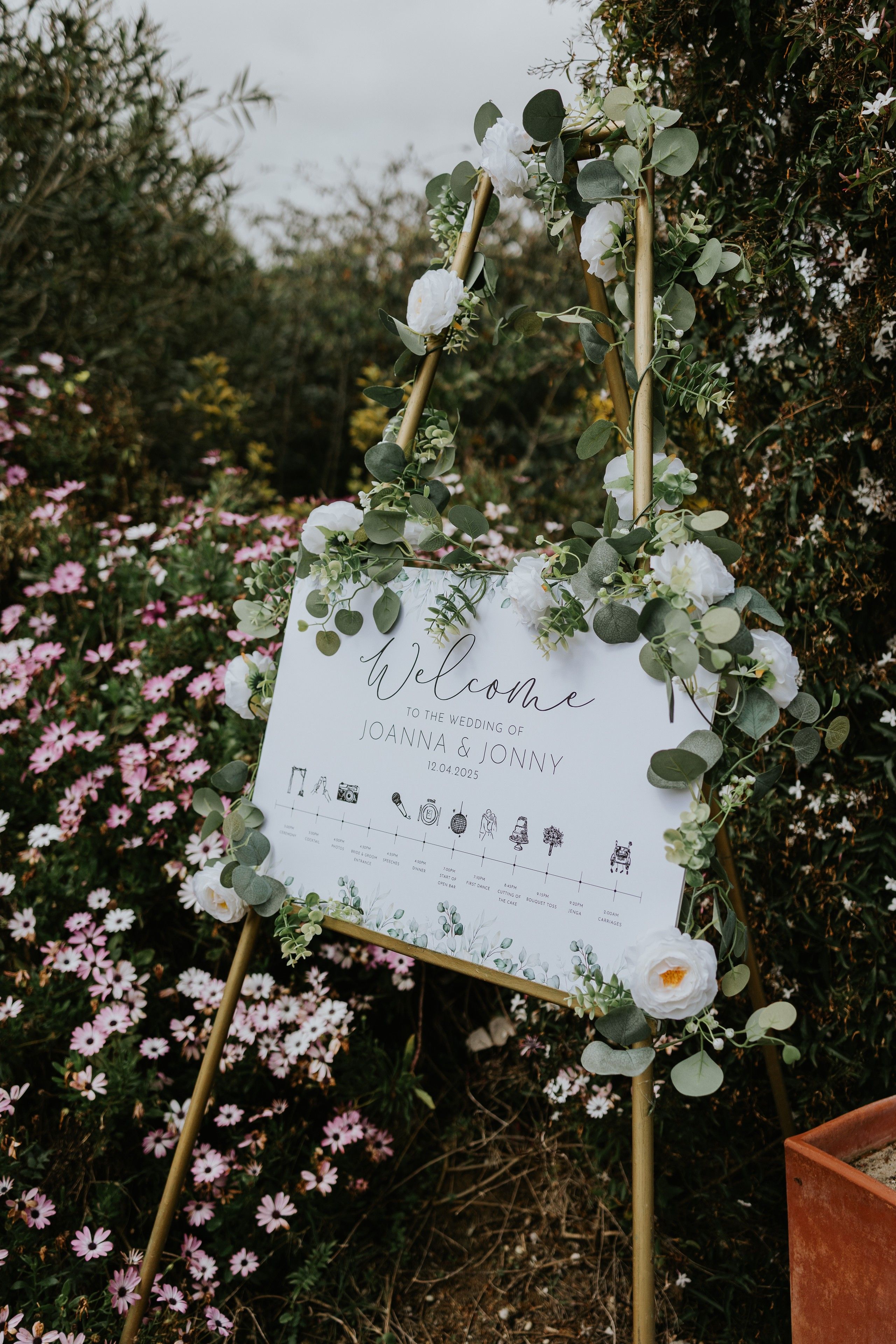 A white wooden wedding board with the word welcome in a stand surrounded by greenery and flowers