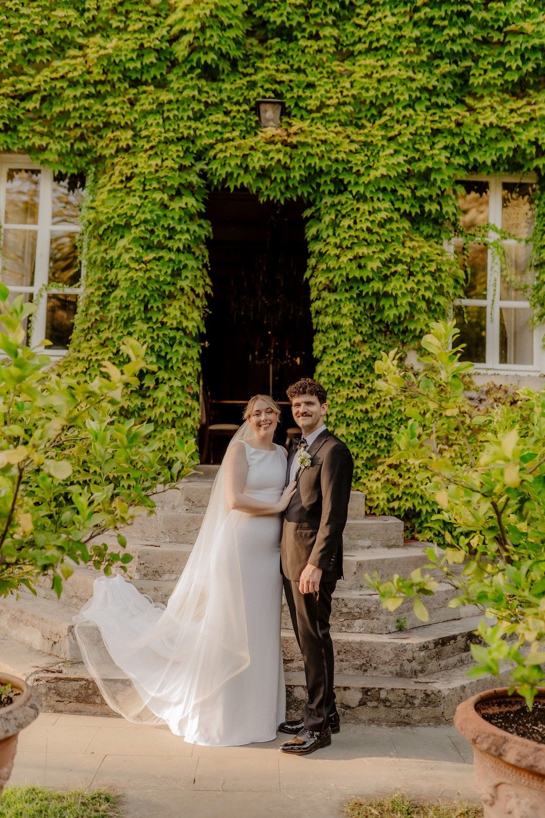Newlyweds look at the camera with the villa's facade covered in greenery in the background during their Tuscan micro wedding