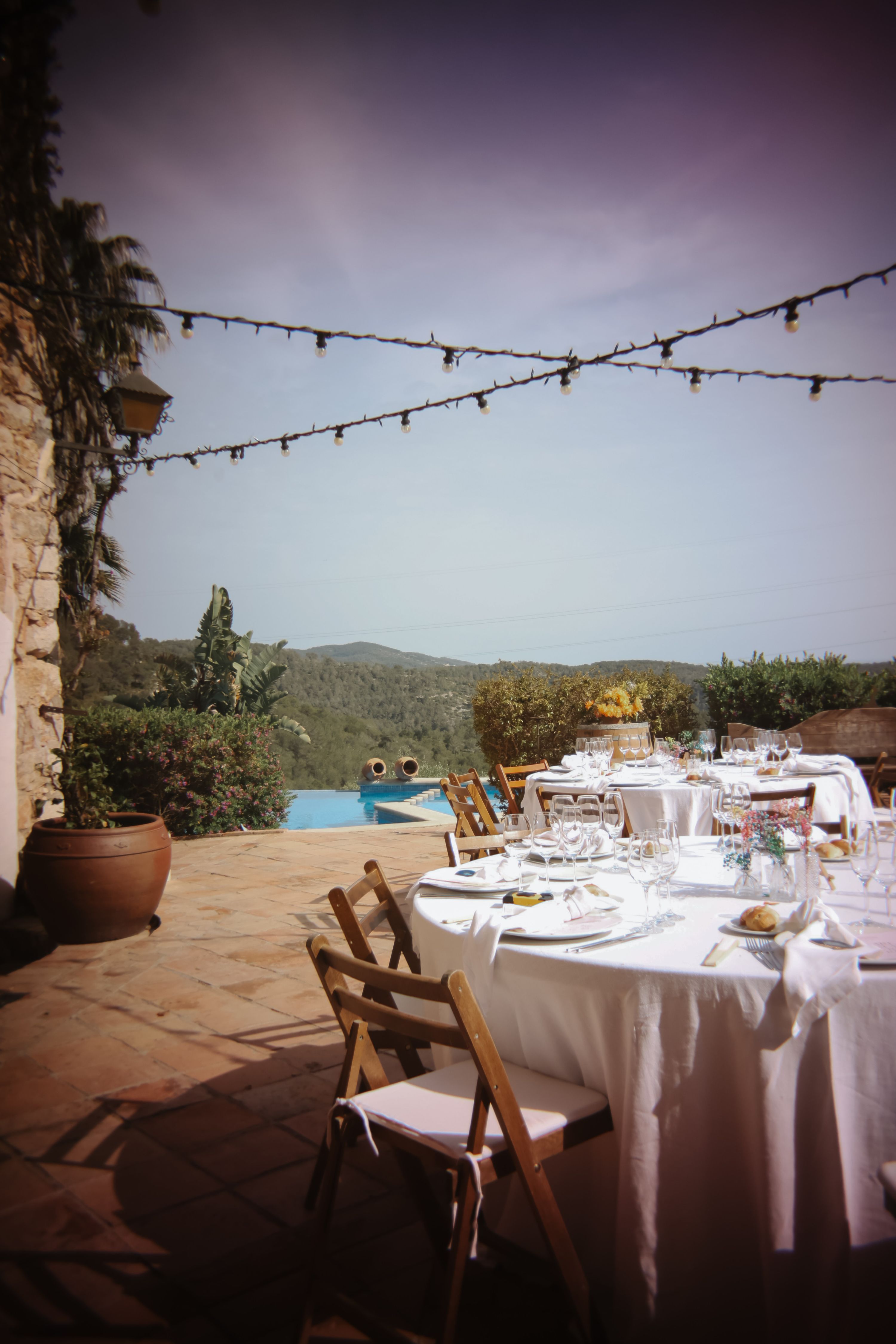 Round tables with wooden chairs arranged outdoors under fairy lights with a pool nearby
