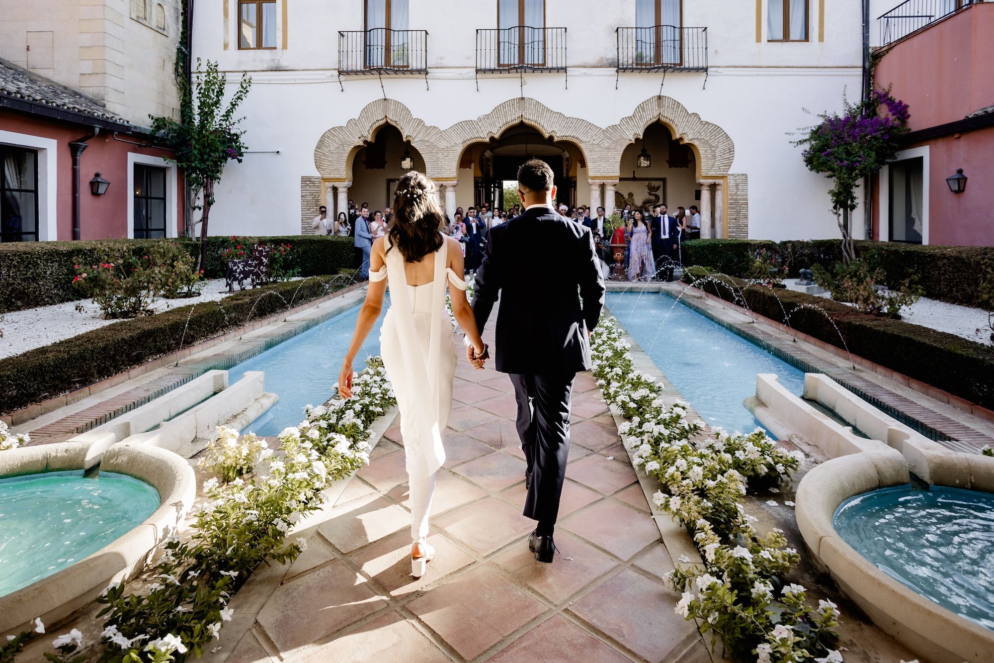 Newlyweds wearing semi-casual outfits walking on a path to their guests waiting on the other side of the building