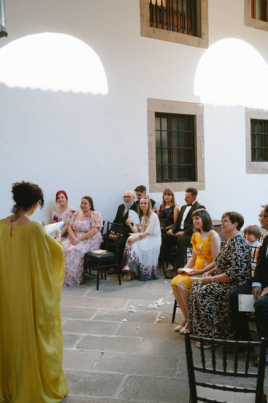 A celebrant in a yellow gown addresses about twenty seated guests under arch shadows in the Parador's cloister gallery.