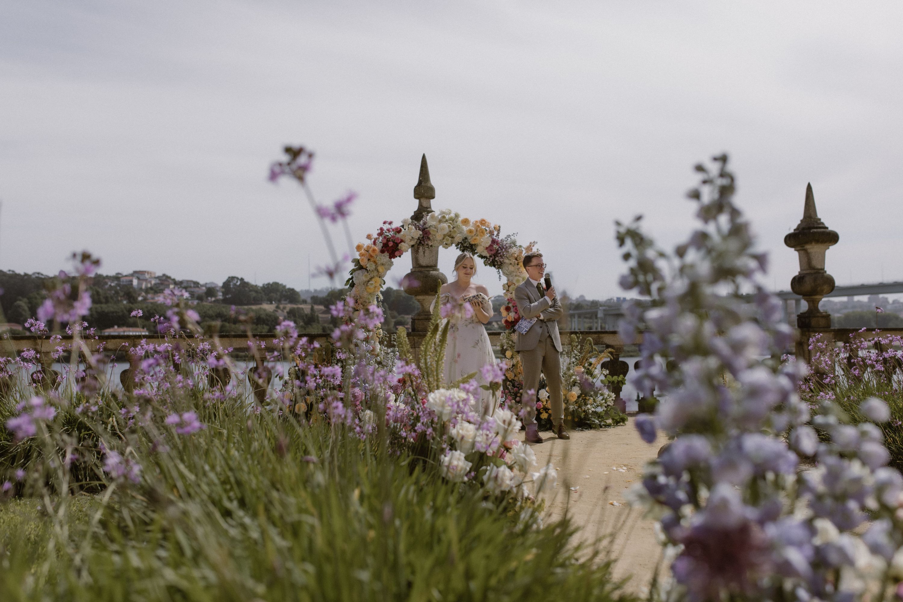Bride and groom exchange vows beneath a floral arch on a riverside terrace, wildflowers in the foreground, at their wedding in Portugal.