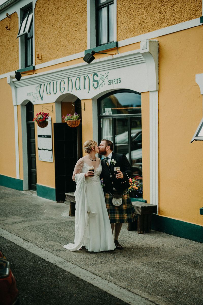 Bride and groom hold a glass of Guinness in front of a yellow Irish pub with the name Vaughan’s written in front of the pub