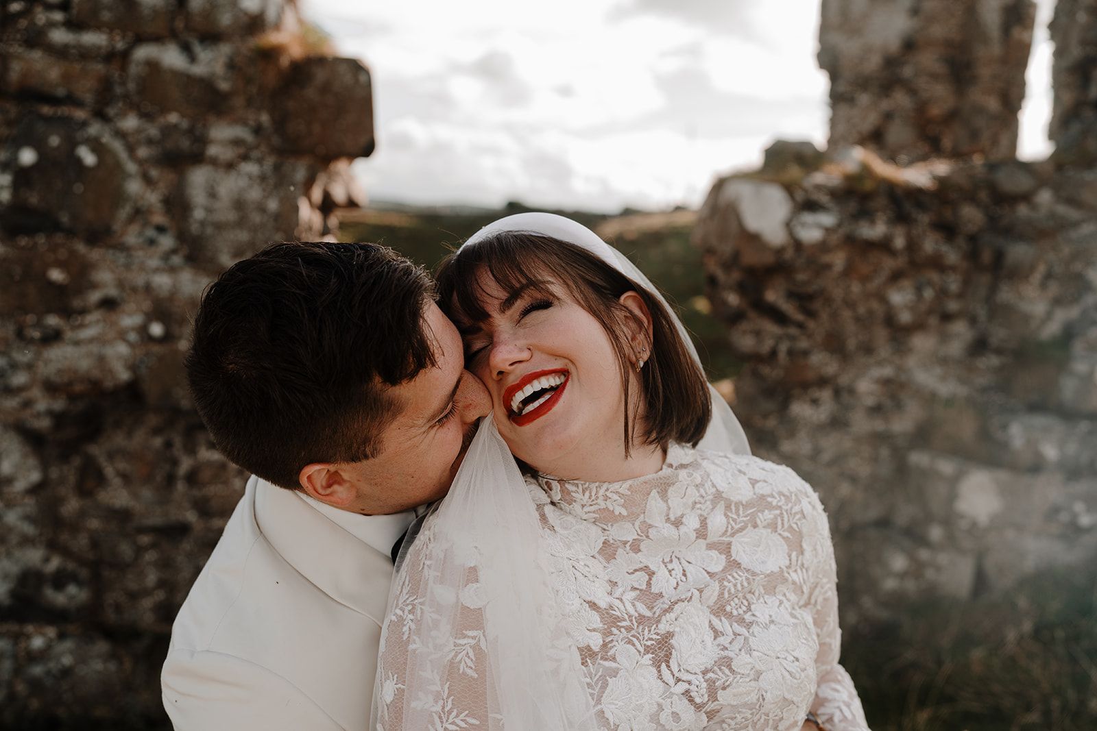 Groom kissing her laughing bride in front of a castle ruin in Northern Ireland