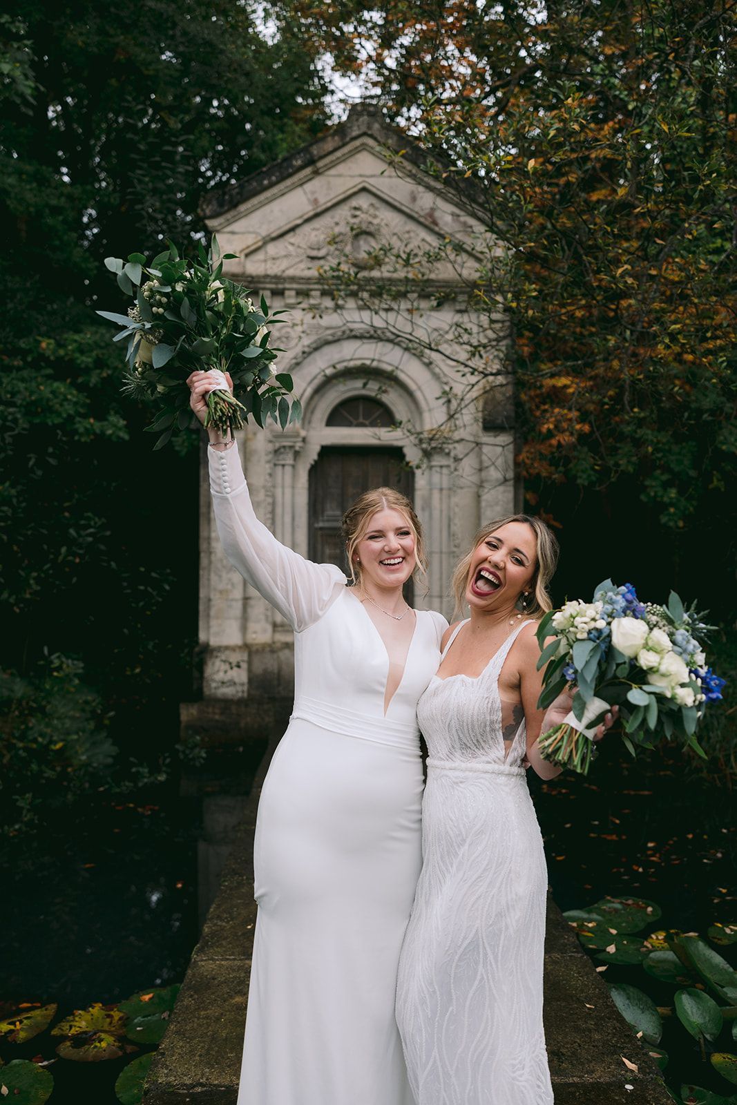 Two brides smiling to the camera while holding their bouquets, with a white stone gazebo and a pond in the background