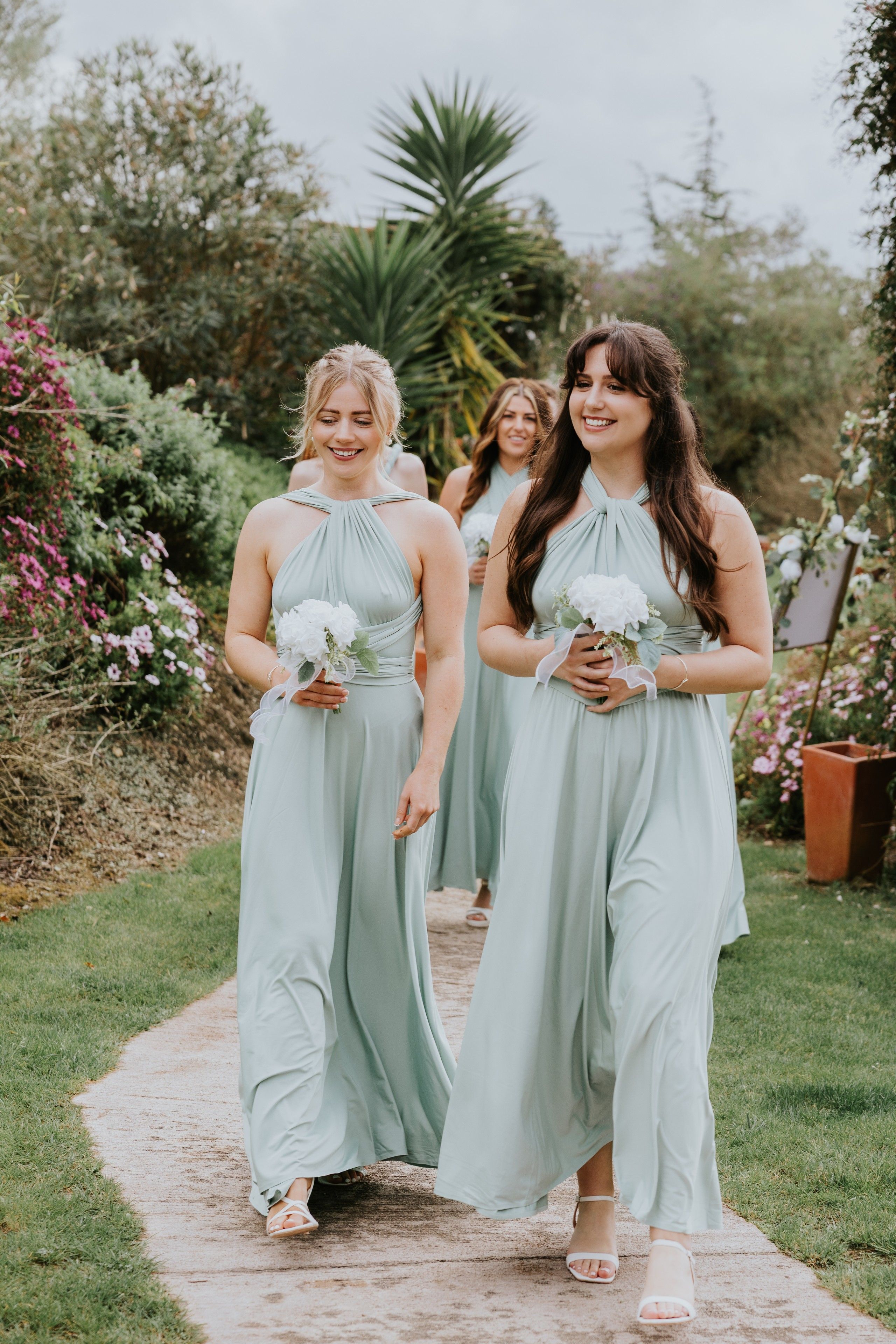 Bridesmaids wearing mint green dresses, holding white mini bouquets walk on a pathway to the ceremony area