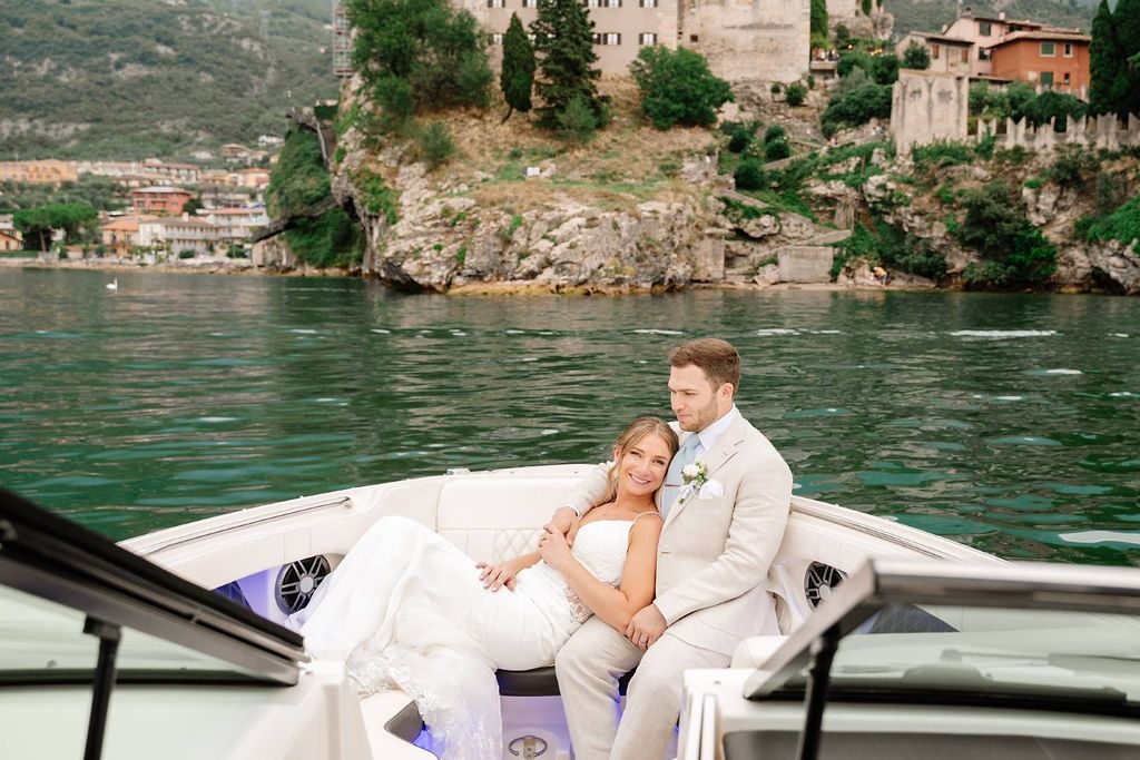 Newlyweds happily ride a boat that sails along Lake Garda while having a photoshoot with a castle in the background