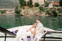 Newlyweds happily ride a boat that sails along Lake Garda while having a photoshoot with a castle in the background