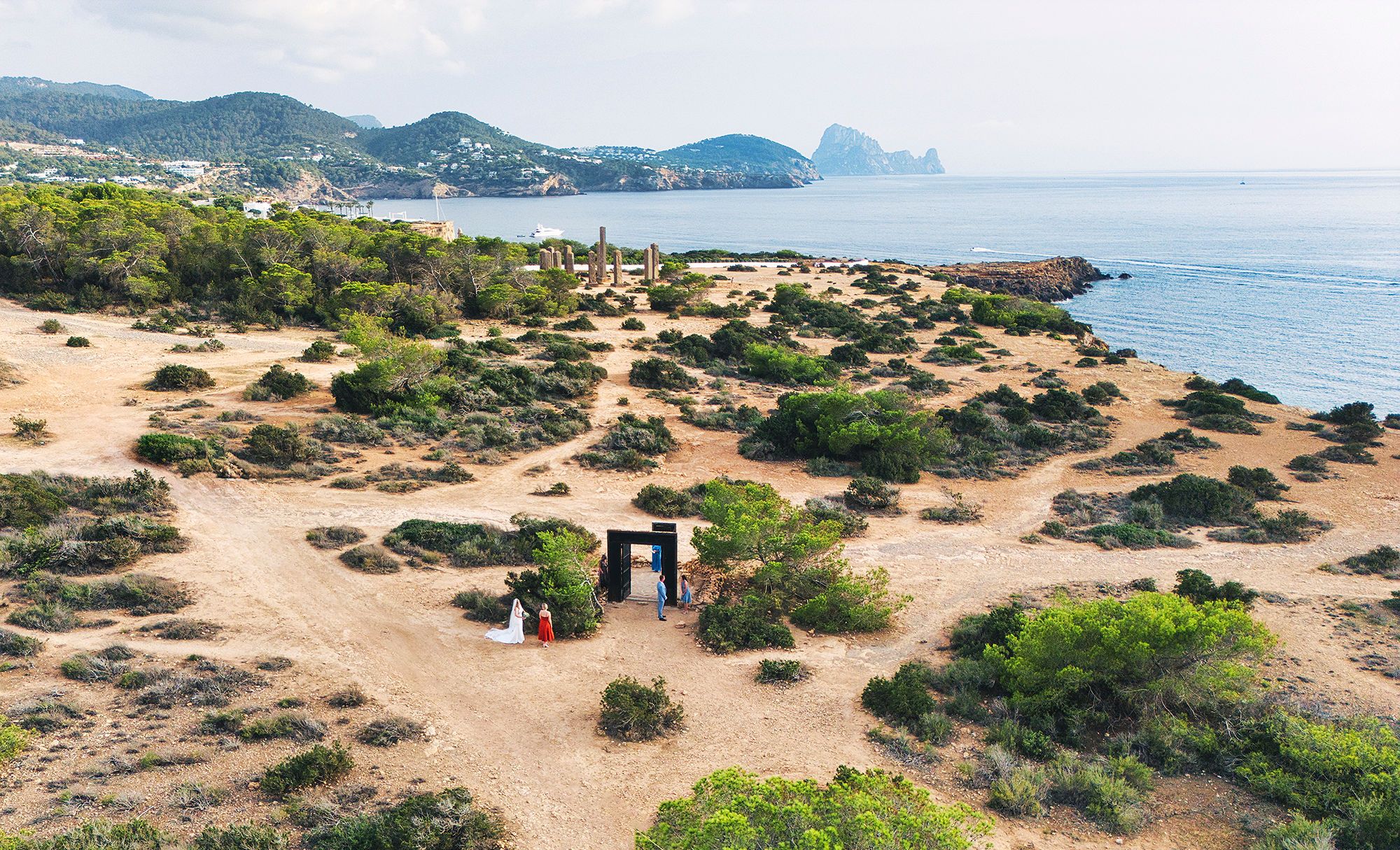 A drone shot of Cala Llentia dotted with greenery in Ibiza during an elopement ceremony with the Balearic sea on the edge