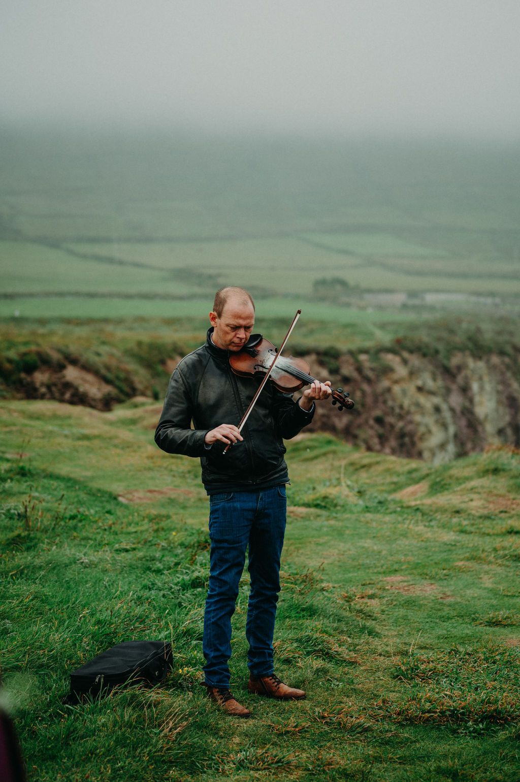 A fiddler in a leather jacket plays a violin on a misty clifftop above Slea Head, his case open on the grass.