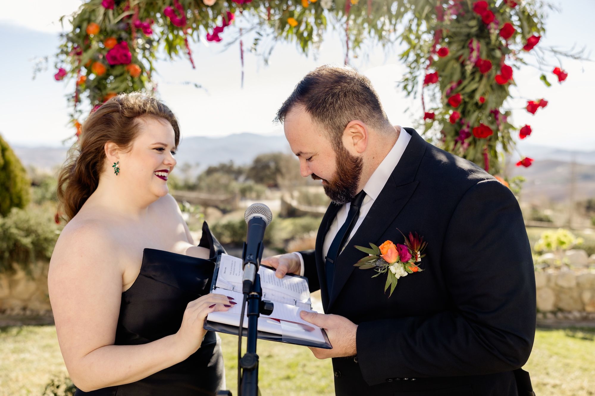 Groom recites vows to his bride on a microphone while the bride smiles during their destination wedding in Spain