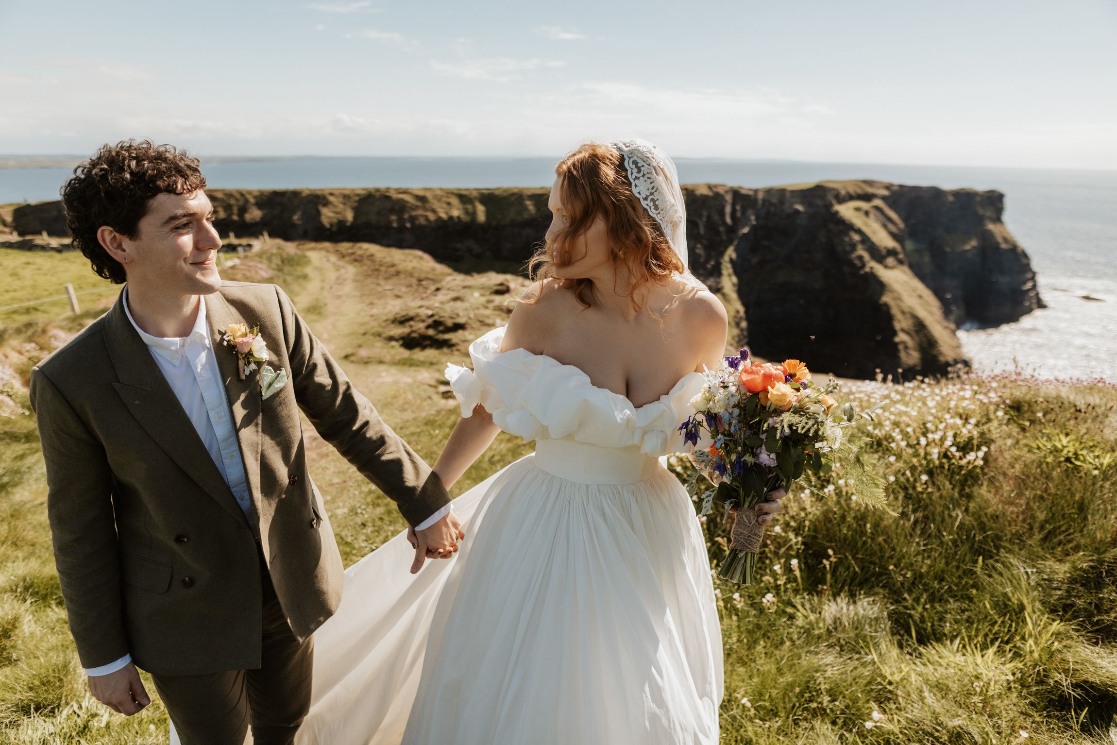 The groom glances back with a smile as his bride carries a wildflower bouquet along dramatic sea cliffs at Hag's Head, Ireland.