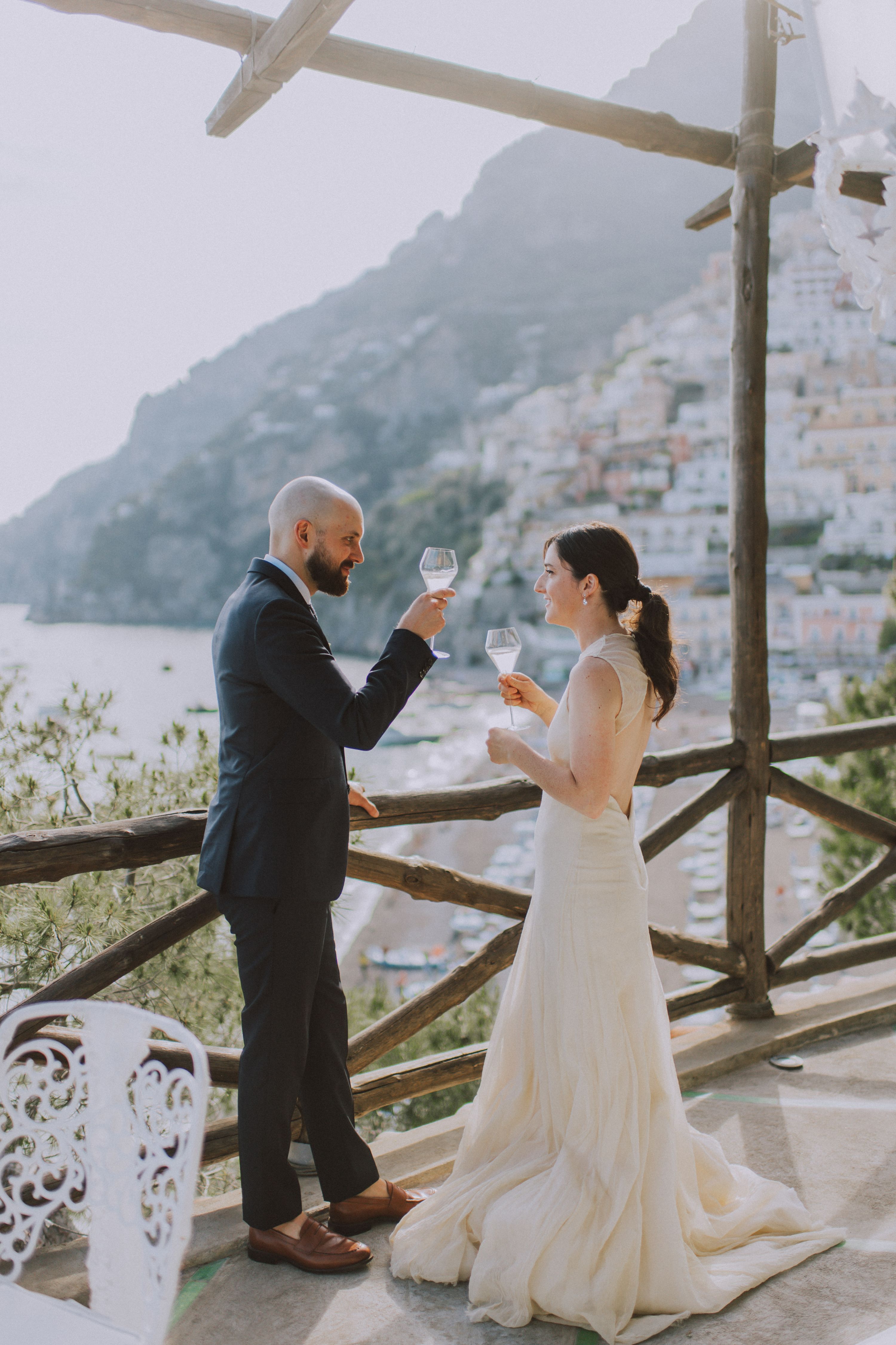 Couple celebrating with wine glasses, while pastel buildings on the hillside of Amalfi and the sea are in the background