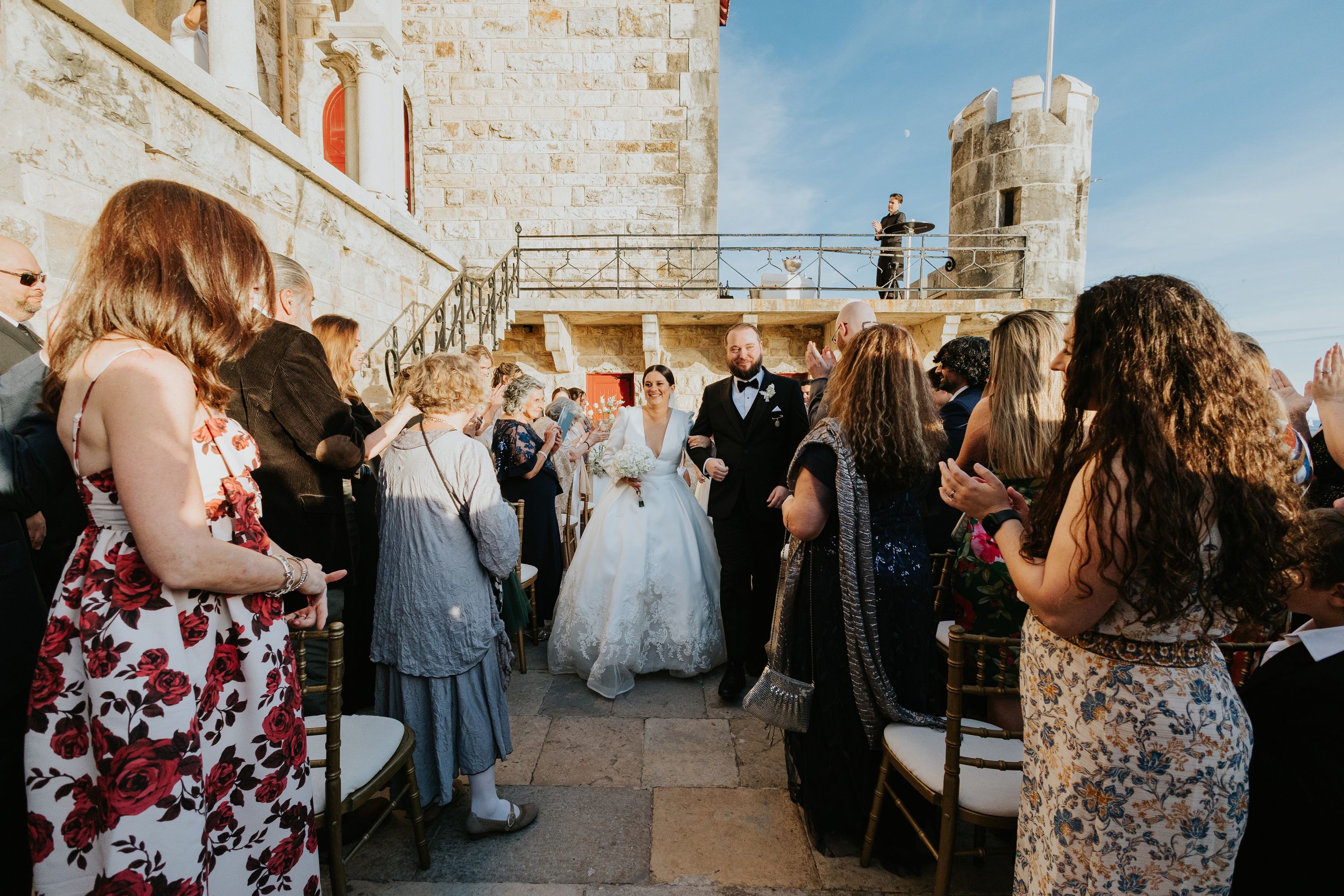 Bride and groom walking down the aisle on top of a terrace of a historic castle in Portugal while their guests look at them in celebration