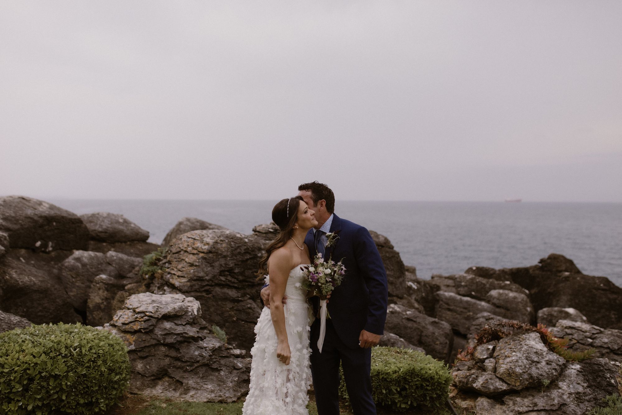 Groom kisses bride on the cheek with rocky coast and sea in the background in Portugal