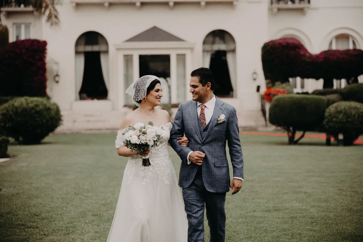 Bride and groom looking at each other walking on a green grassy field with a white building in the background