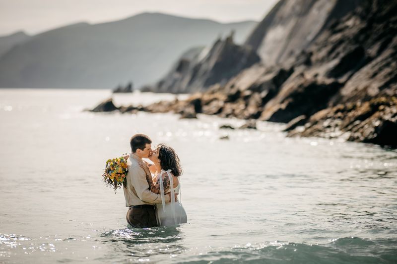 Bride and groom kiss while half-submerged in the ocean with cliff rocks and mountains in the backgrounds