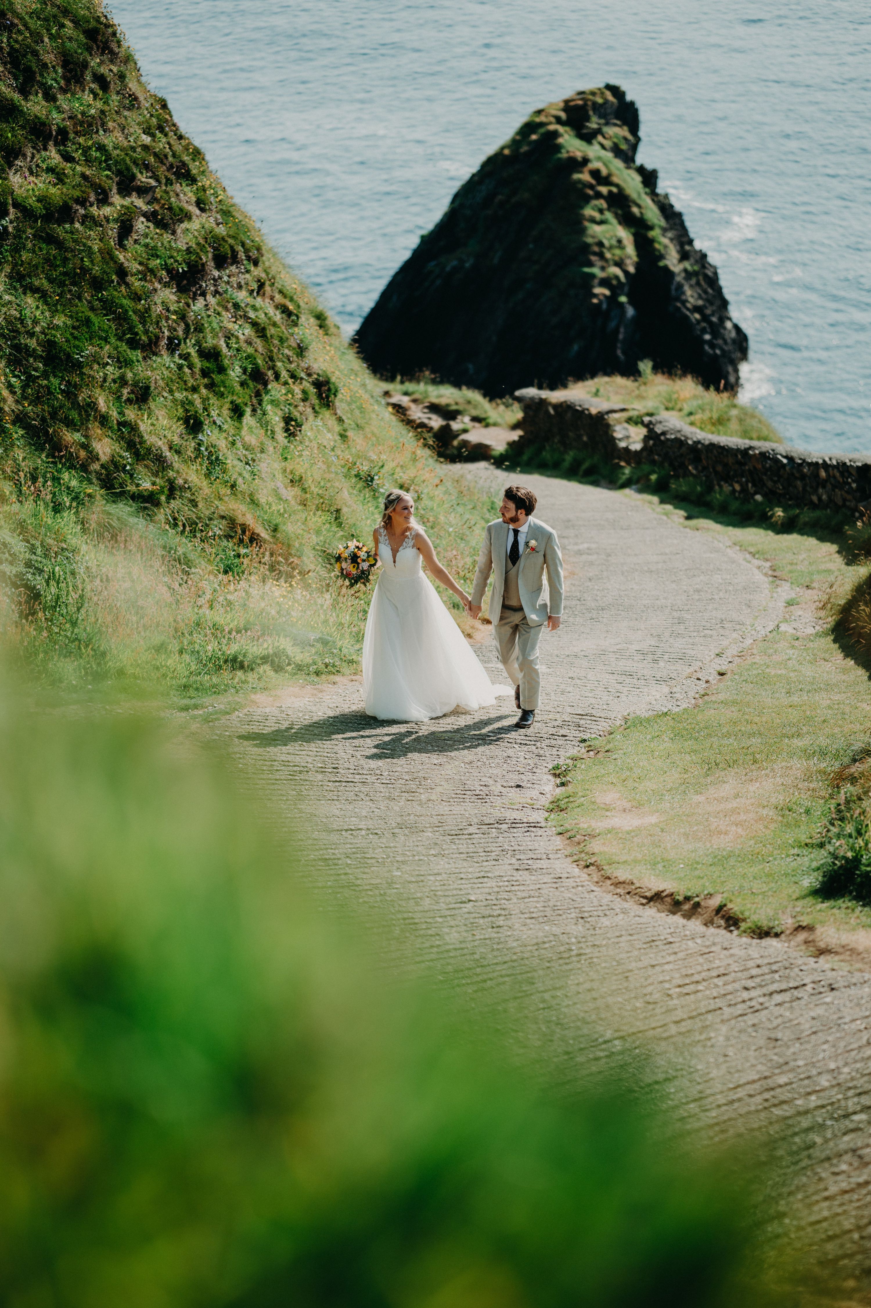 Newlyweds walk hand in hand up the curved concrete path from Dunquin Pier, a sea stack rising from the water behind them.