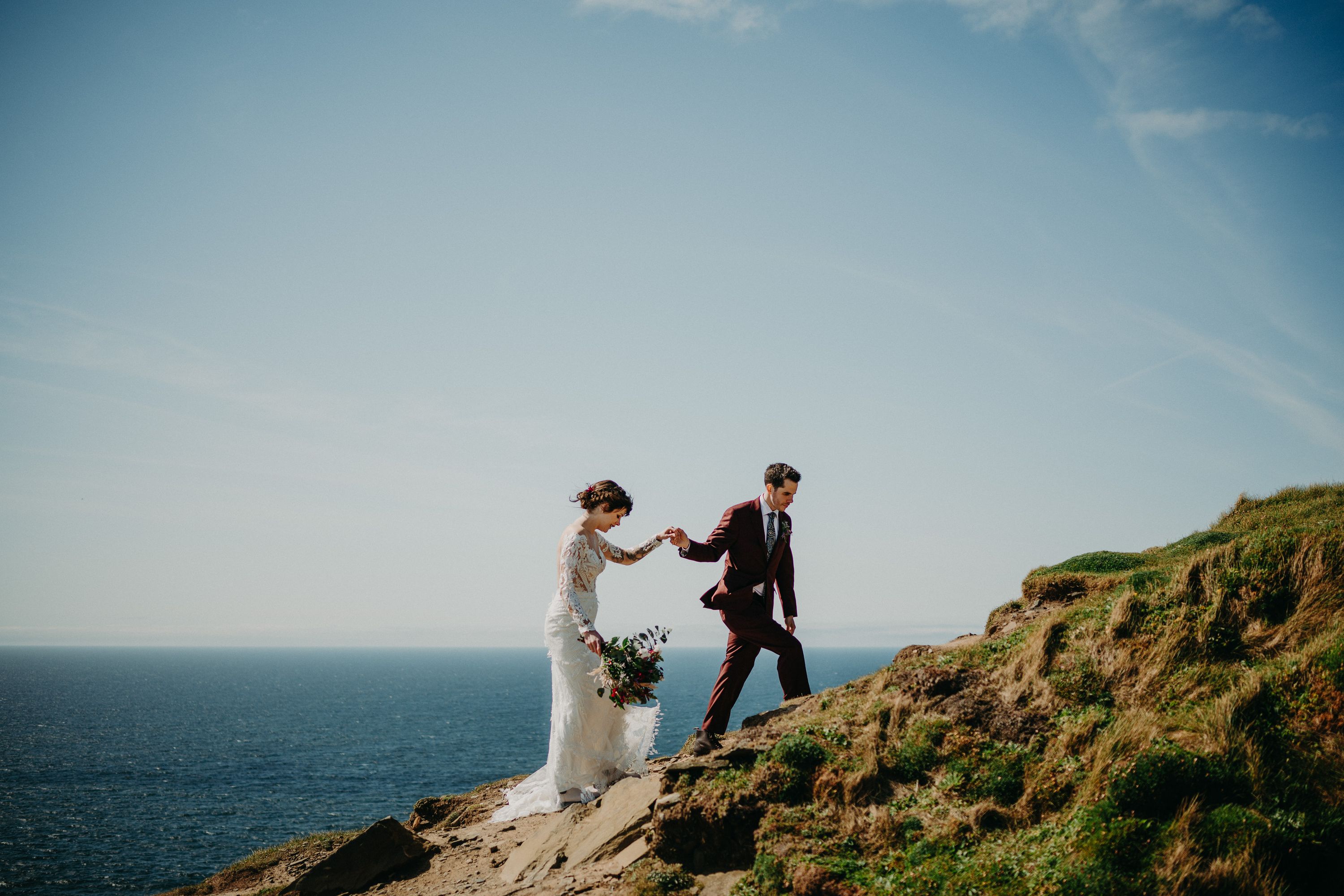 A bride and groom climbing up a cliff with the blue sky and ocean behind them after their small wedding in Ireland