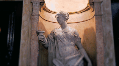A warmly lit classical statue holding an urn stands in a wall niche inside a grand Portuguese palace.