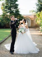 Bride and groom wearing wedding attire, drinking champagne in an open space with lush greenery behind them