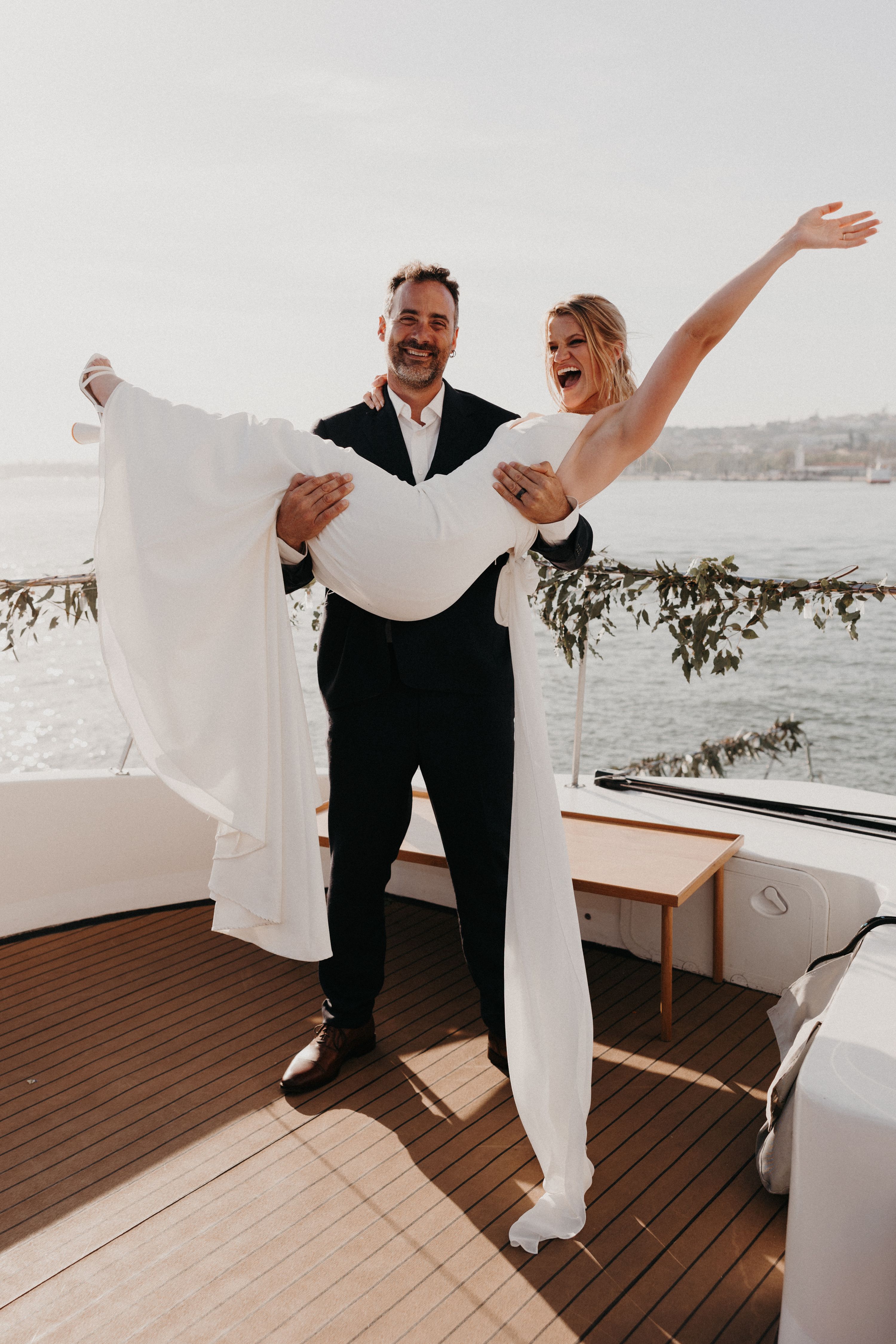 Groom carries his bride ecstatically on a boat sailing across Tagus River during their small wedding in Portugal