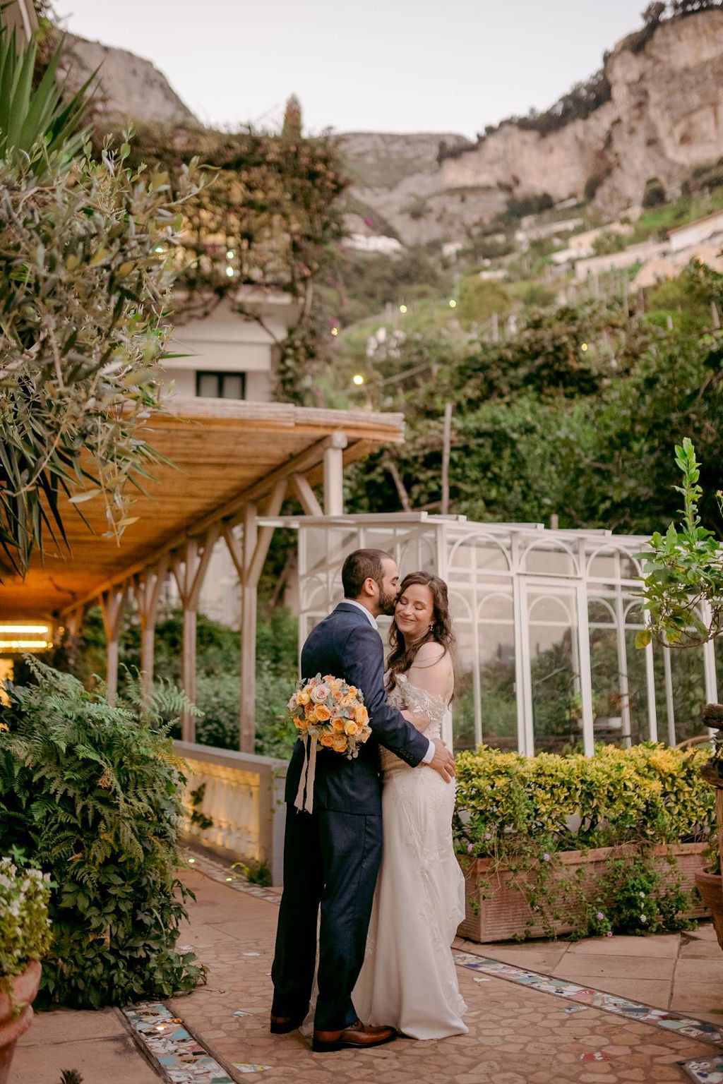 Groom kisses his bride who smiles and holds a bouquet during the photoshoot of their elopement in Italy at the Amalfi Coast