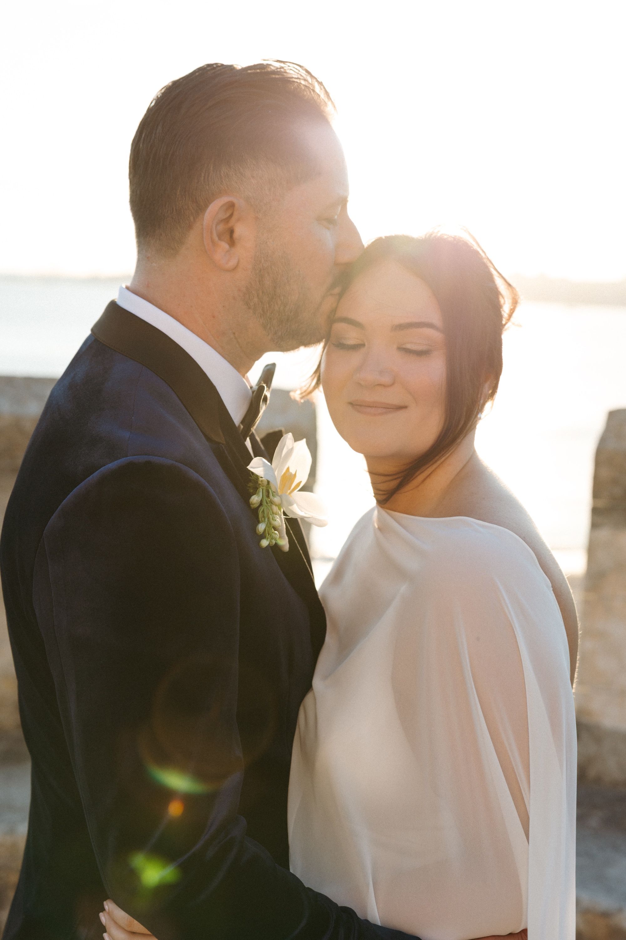 Groom kisses his bride on the temple with the sunset behind them during the photoshoot of their Wedding guests gathered in the castle's balcony for the drinks reception of a small wedding in Portugal