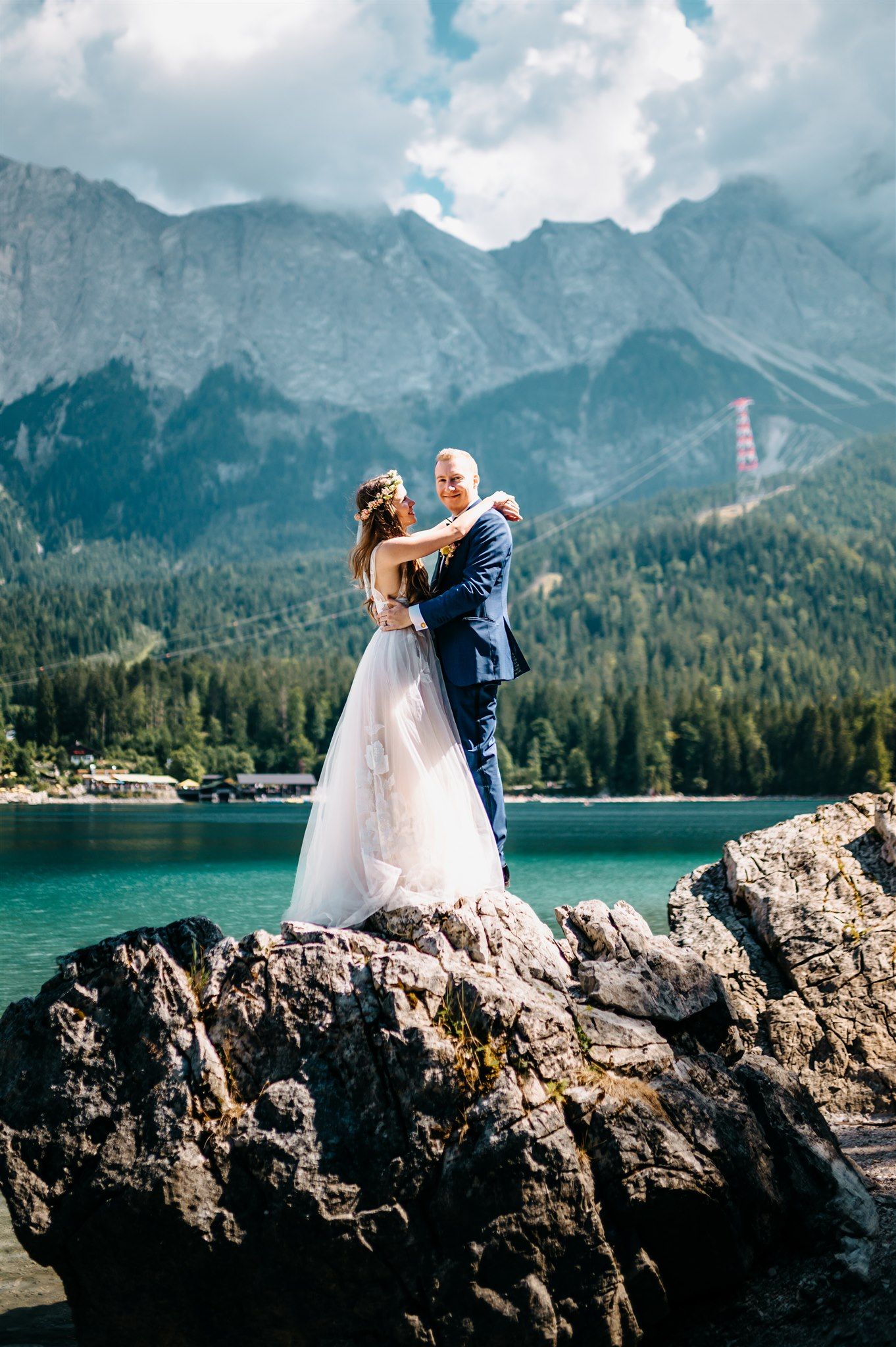 A bride and groom standing on a rock beside a lake with the mountains behind for their micro wedding in Germany