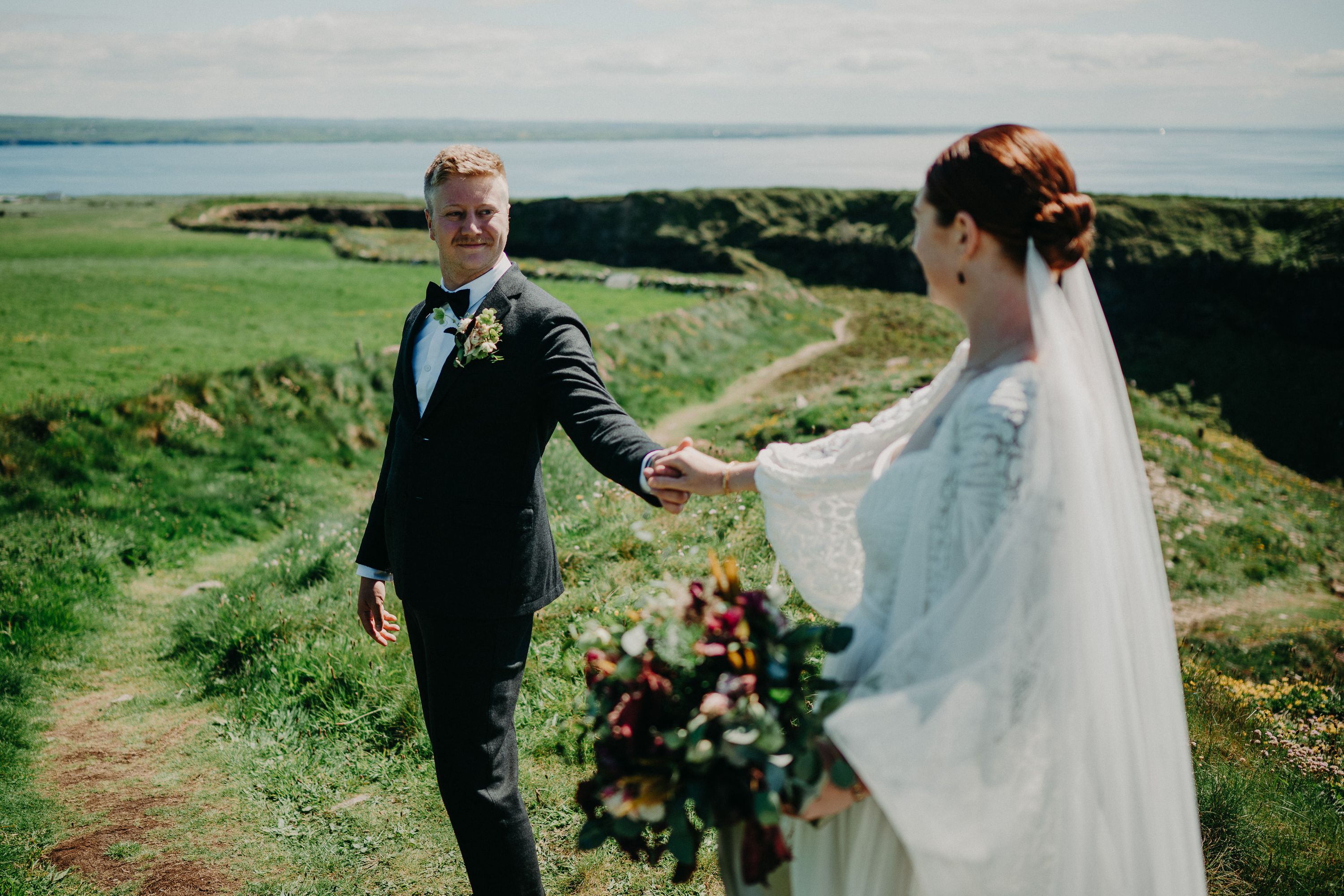 A groom holding his bride's hand along a grassy path after their small wedding in Ireland