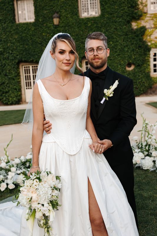 Bride and groom holding each other and looking at the camera with a building covered in greenery in the background