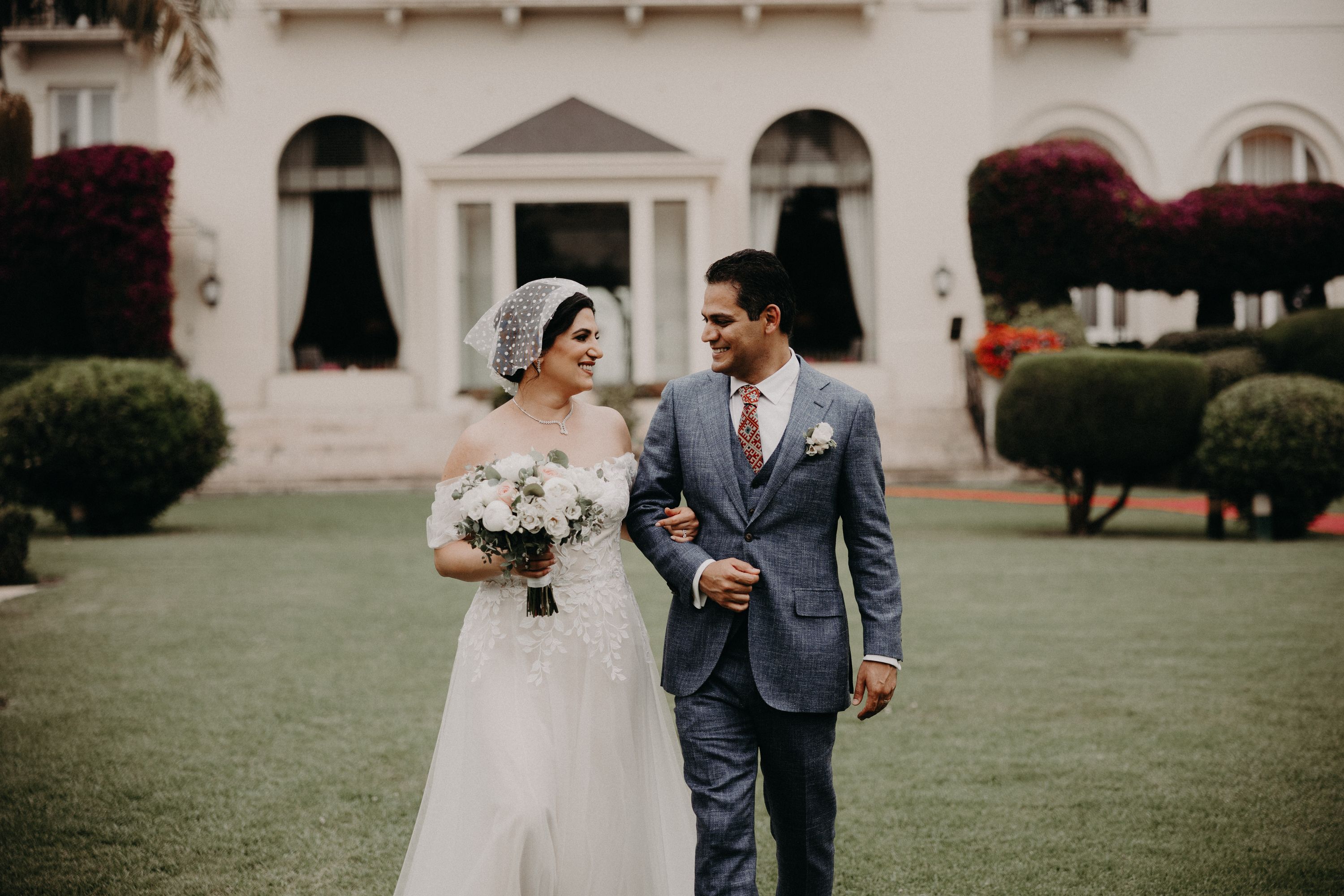 Bride and groom looking at each other while walking on a grassy area with a white hotel building at the back