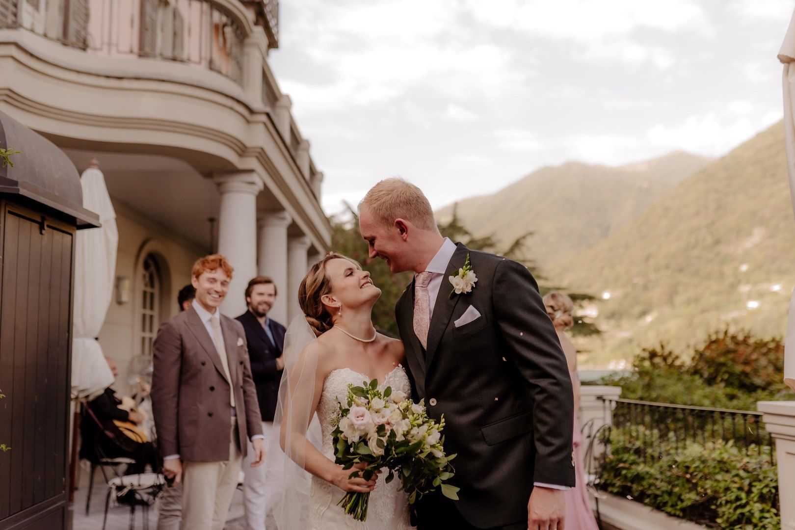 Newlyweds kissing with a few guests smiling, as well as Lake Como and Italian rolling hills in the background