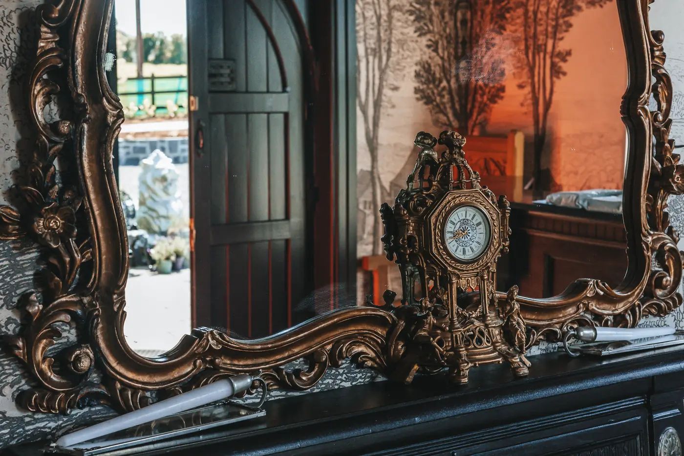 Gilt mantel clock and ornate mirror reflecting the castle's open arched door onto the sunlit forecourt.
