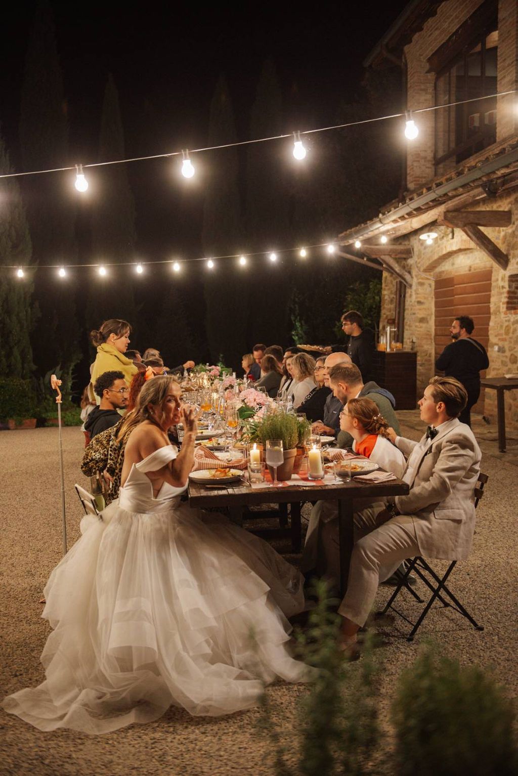 Bride and groom seated in a long table with their friends and family under string lights during their small wedding in Italy