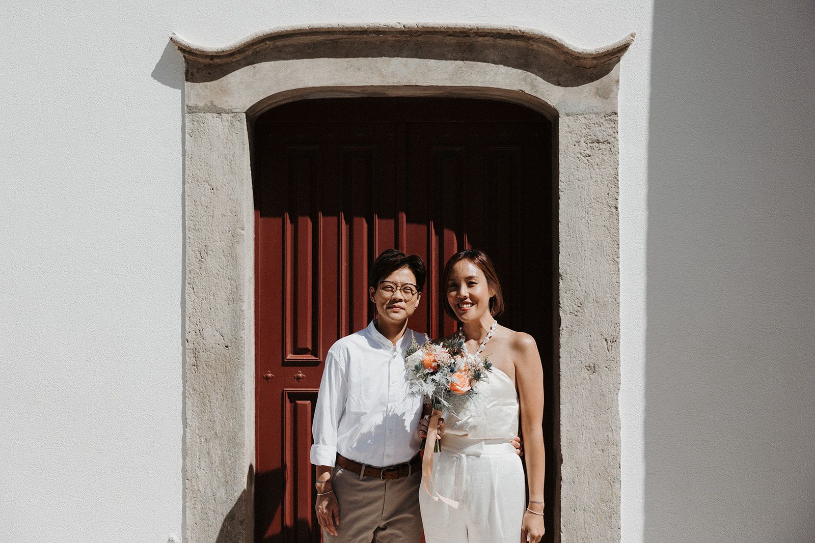 Bride and groom in casual attire having a photoshoot with a red wooden door in the background during their destination elopement in Portugal