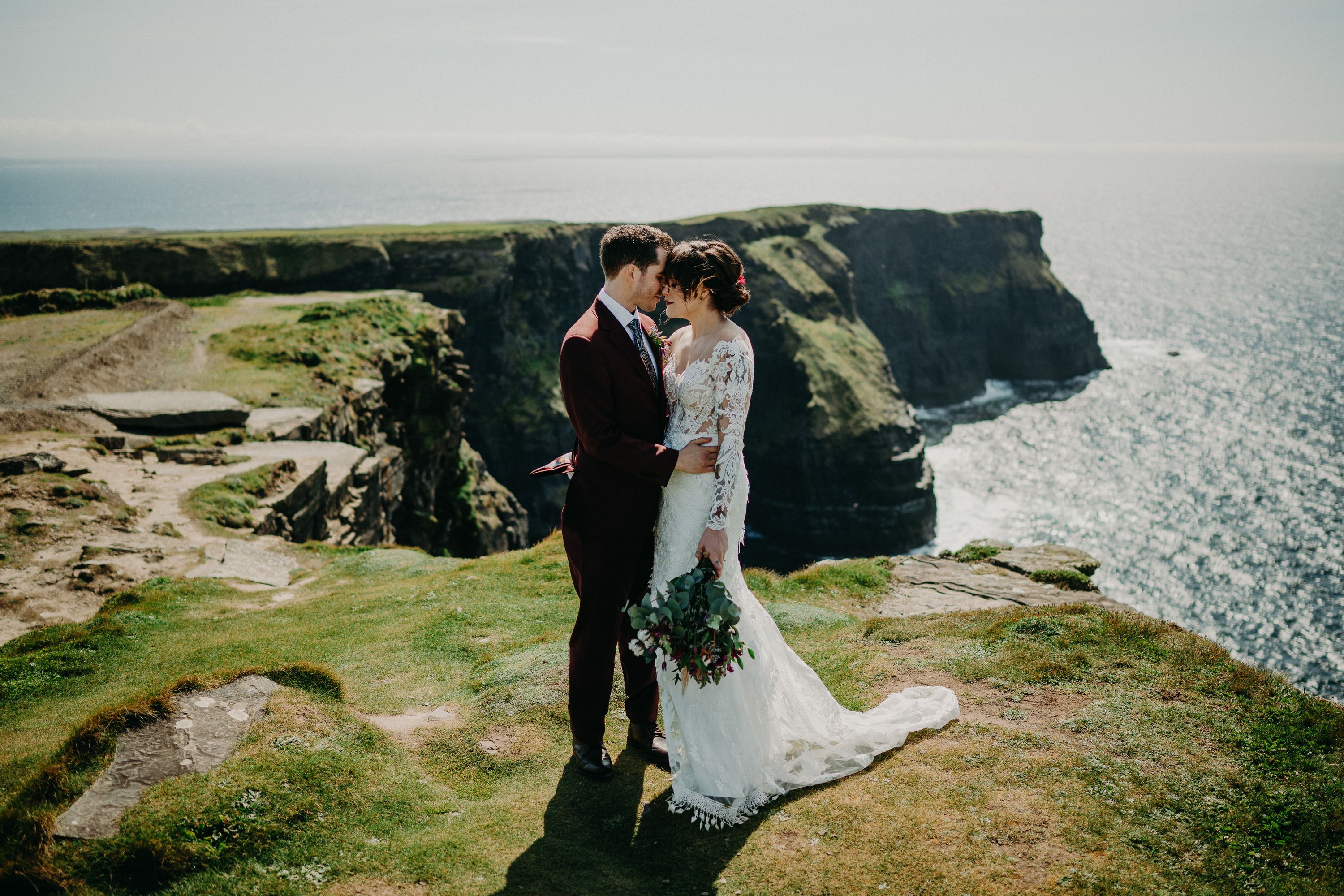 A bride hugs his groom as they stand on top of a majestic cliff with a lovely view of the sea after their small wedding in Ireland