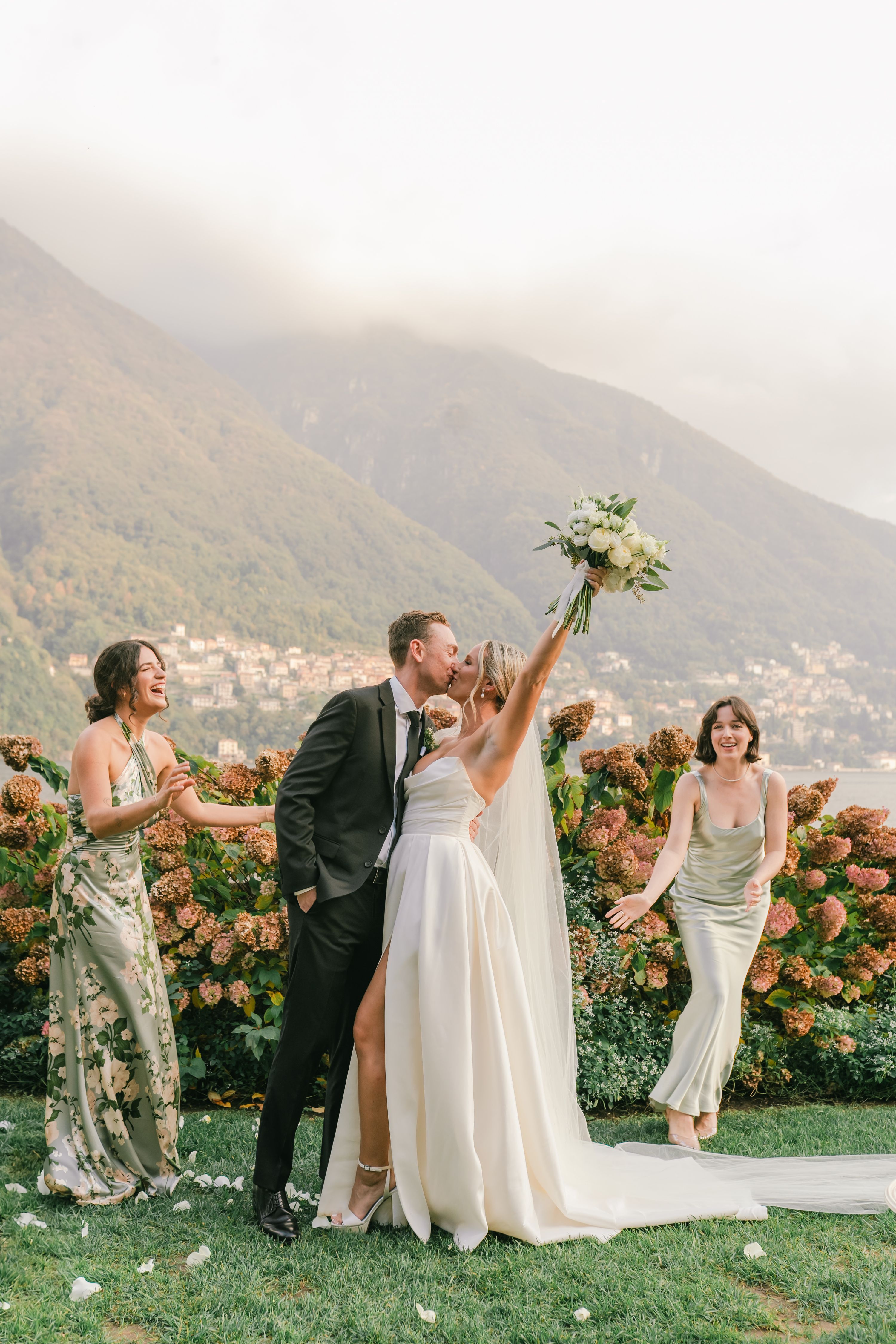 Newlyweds kiss with bridesmaids cheering, Lake Como and Italian hills in the background as the couple renew vows in Italy