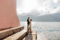 A view of Lake Maggiore with a couple standing on the ledge and a portion of a large orange building on the left