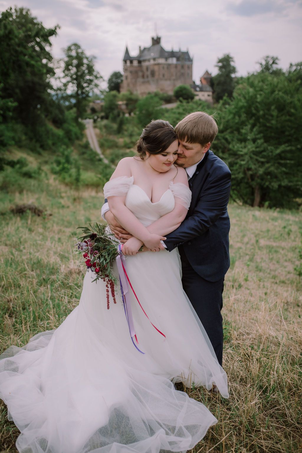 A groom hugging his bride with a castle surrounded by trees during their intimate wedding in Germany