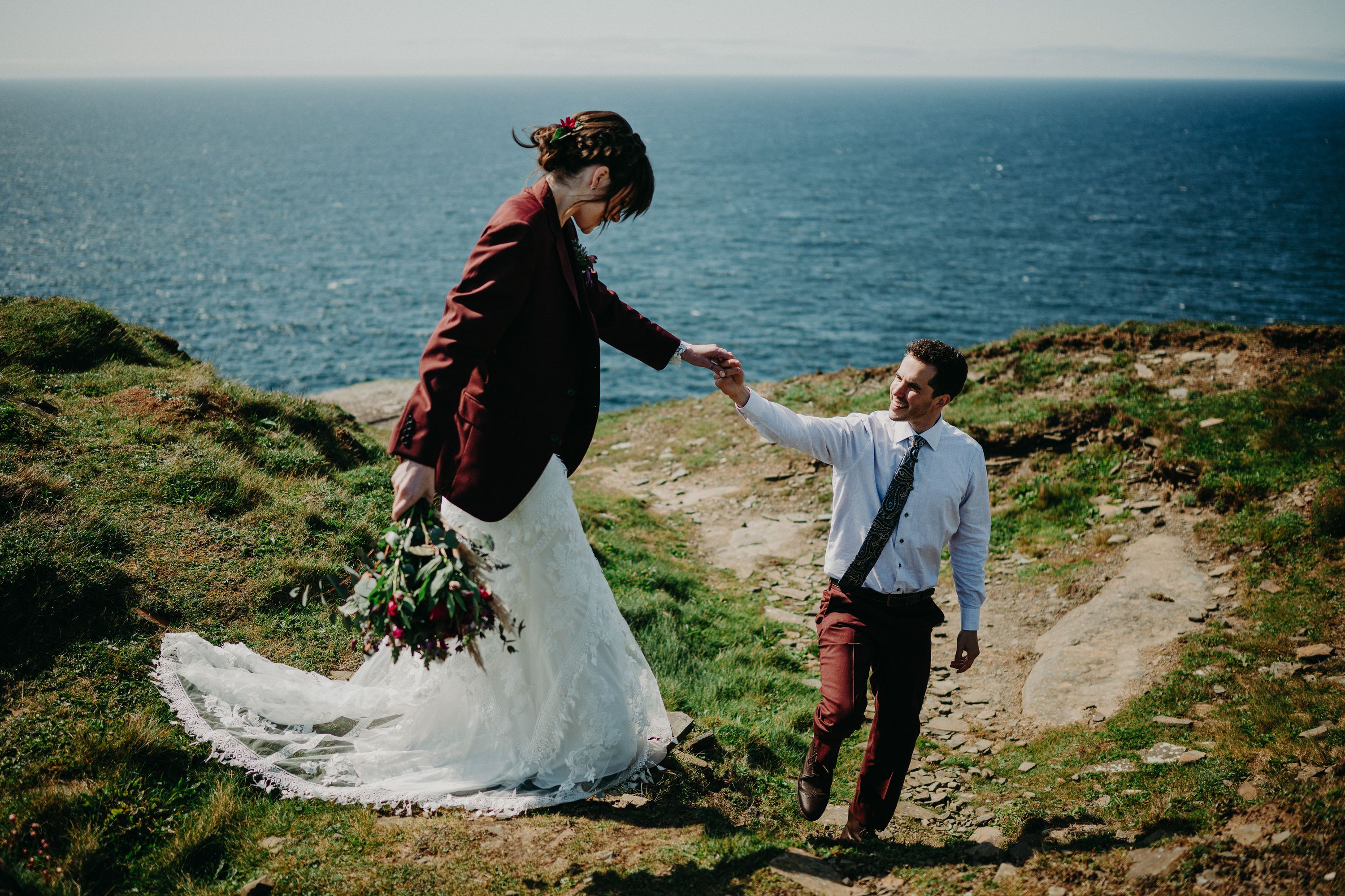 A groom helping his bride walk down the cliff with the ocean behind them after their intimate wedding in Ireland