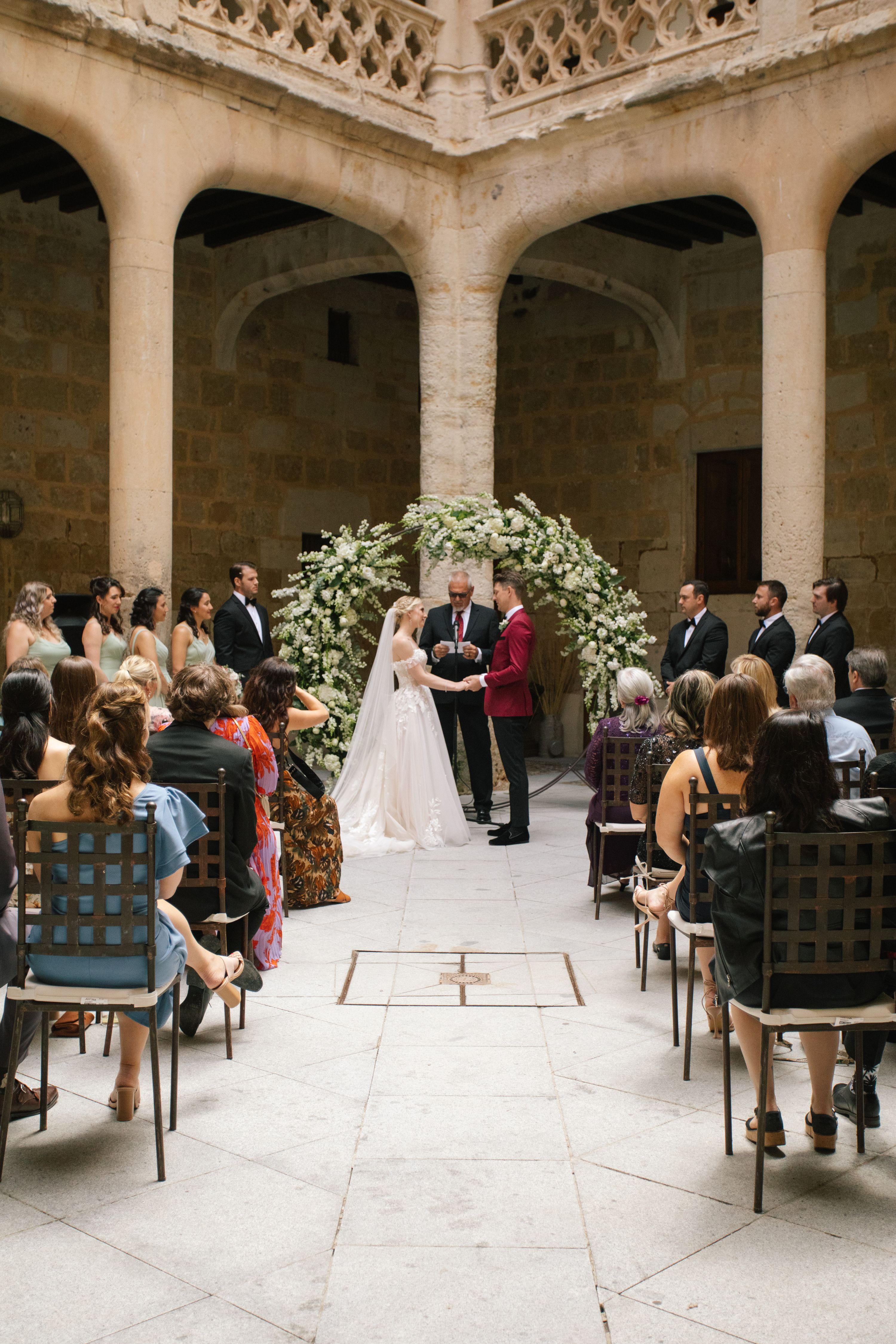 A ceremony in a courtyard with a floral arch, the bride and groom, and entourage in front during a destination wedding in Spain