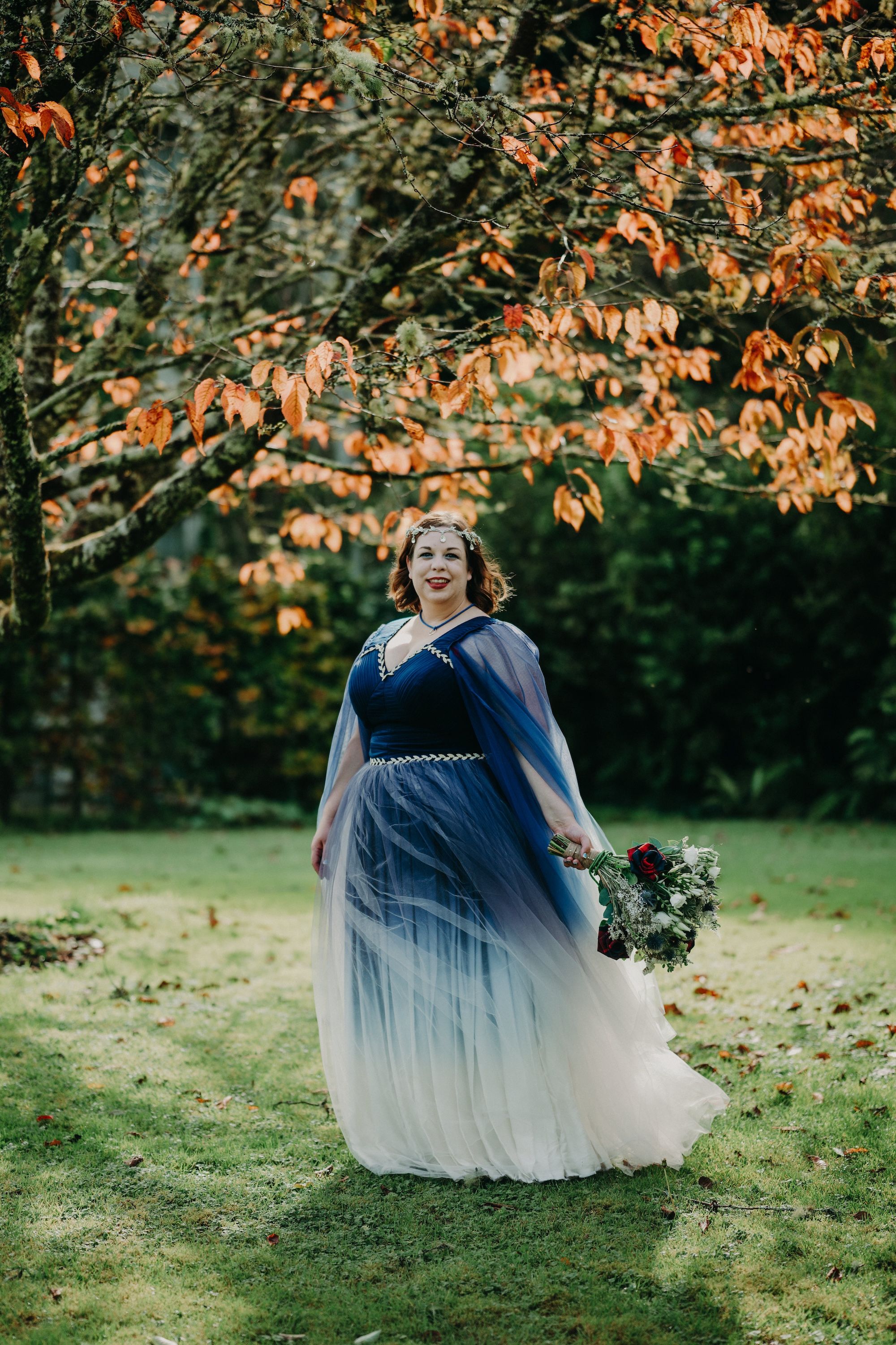 Bride under an autumn-colored tree wearing a blue and white ombre bridal gown, holds a bouquet, on her elopement in Ireland
